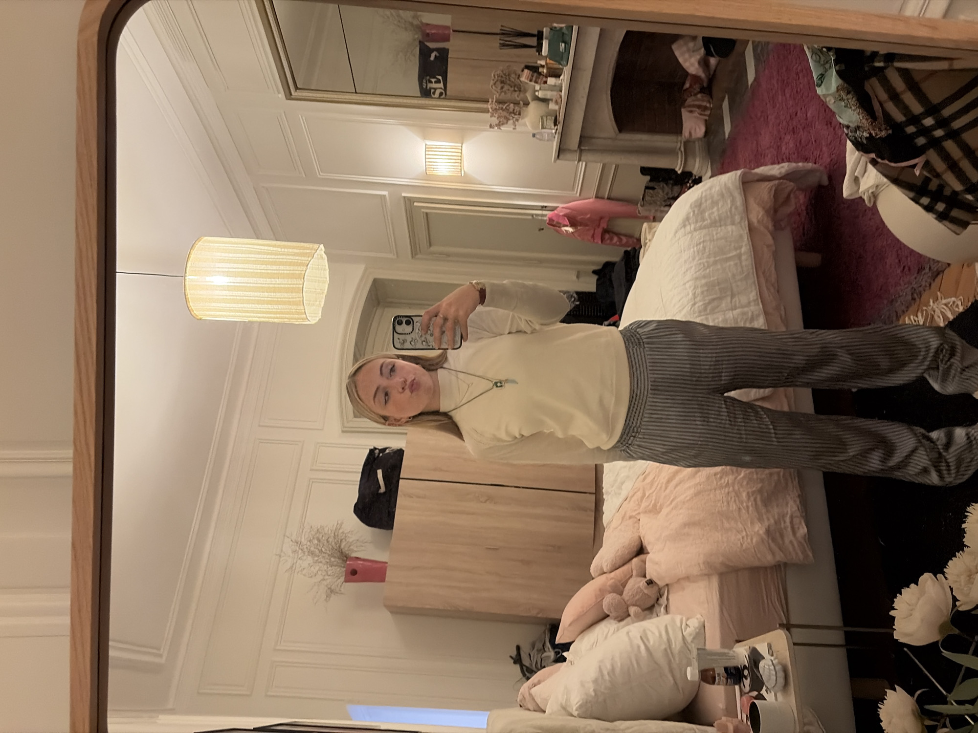 The image displays a young, fair-haired female, captured from the waist up in a mirror selfie within what appears to be a residential bedroom in Paris, France. The room's architecture, including ornate ceiling moldings, intricate wall paneling, and a classical marble fireplace mantel, is characteristic of traditional Parisian apartments.
The female subject wears a cream-colored long-sleeve turtleneck top and grey pinstriped trousers. She exhibits a pouting facial expression and holds a smartphone, equipped with a white patterned case, in her right hand, partially obscuring her face. A pendant necklace is visible around her neck.
The bedroom is furnished with a large bed on the left, featuring a white duvet, light pink sheets, several pillows (white and pink), and a small light brown teddy bear nestled among them. A small, light-colored side table adjacent to the bed holds a dark mug, a small white dish, and a brown bottle. To the left of the subject, a light-wood wardrobe is topped with a pink vase containing dried plant material and a dark bag. To the right, a white marble fireplace mantel is surmounted by a large mirror, which reflects portions of the room and an inverted "SE" text. The mantelpiece itself displays an assortment of bottles, containers, and dried flowers in a white vase. A pink jacket hangs on the wall or a door near the fireplace.
The room is illuminated by a central cylindrical pendant light fixture and a wall sconce with a similar textured shade. The floor is predominantly covered by a dark pink shag rug, with wooden flooring visible closer to the fireplace. In the lower right foreground, a light-colored stool or ottoman is visible, draped with folded clothes, including a plaid-patterned item. White artificial flowers are positioned in the lower left foreground.
