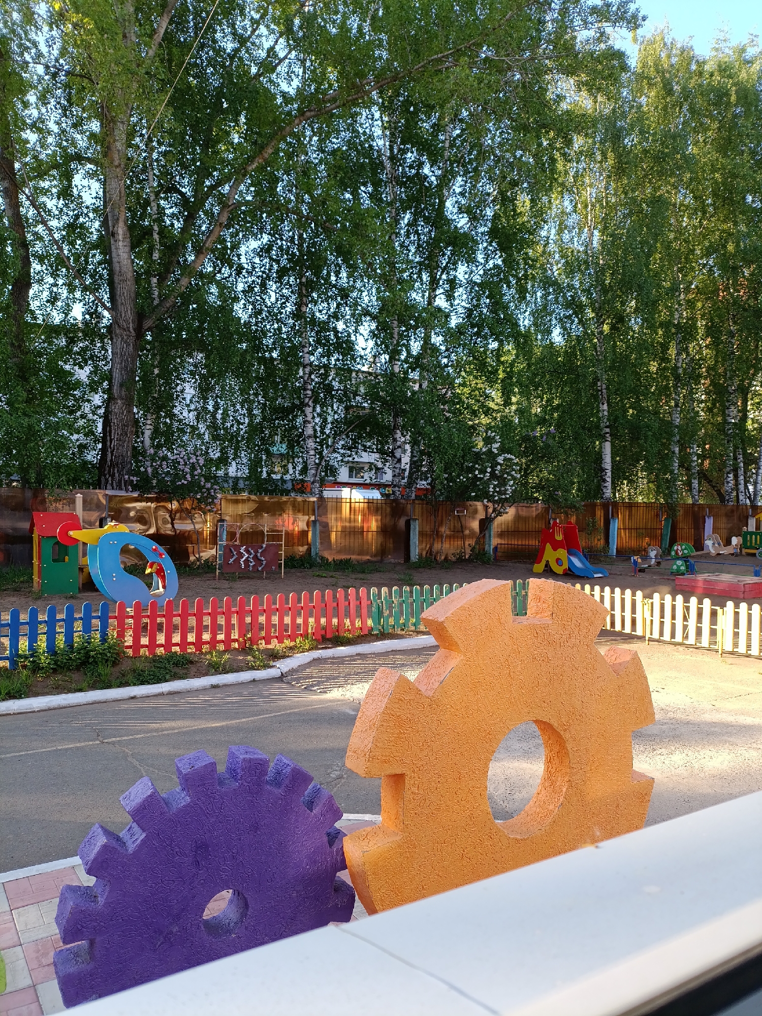 The image depicts a playground in Chaikovsky, Russia. In the foreground, two large, decorative gears, one purple and one orange, are prominently displayed. The playground features various play structures including a colorful climbing structure with a slide, a climbing frame with animal symbols, a red and blue slide, and a sandbox with assorted toys. The playground area is bordered by a wooden fence of various colors. Behind the playground, a wooden fence separates the playground from a grassy area with numerous tall birch trees. A building is visible in the background through the trees. The playground surface is a mix of asphalt and sand. The lighting suggests it is daytime. No people are visible.