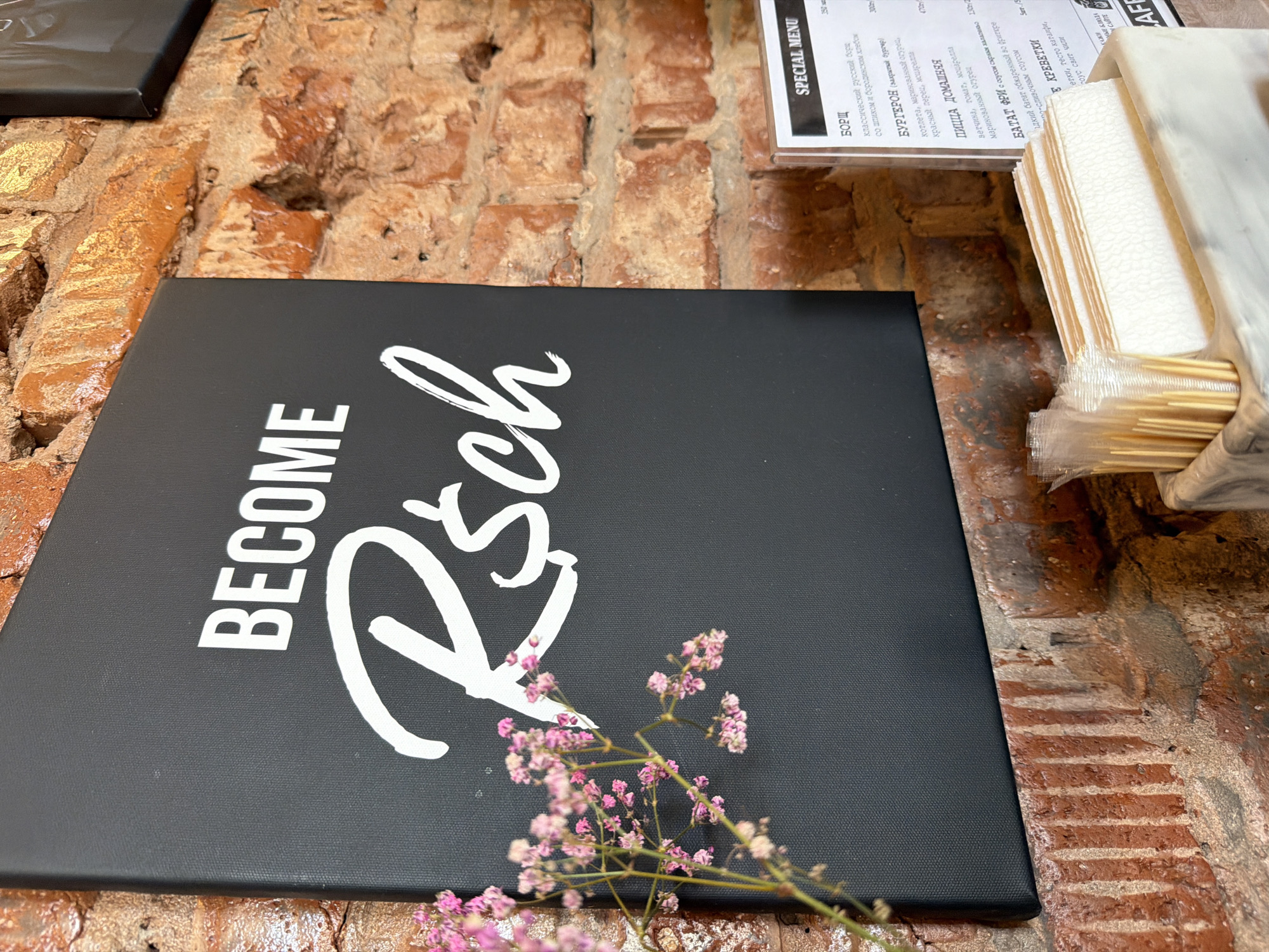 The image captures an interior view of an establishment in the city of Chaikovsky, Russia. A black rectangular canvas sign, featuring white typography that reads "BECOME" in a sans-serif capital font and "R$ch" in a script font with a dollar sign replacing the 'i', is mounted on an exposed, rough brick wall. The bricks exhibit varied shades of reddish-brown and orange, with visible mortar lines. In the left foreground, a delicate branch of small pink flowers is partially visible and slightly out of focus. To the right, a clear plastic menu holder displays a "SPECIAL MENU" written in Cyrillic, listing items such as Borscht, Burgers, Homemade Pizza, Sweet Potato Fries, and Shrimp. Beneath the menu, a container holds multiple stacks of white, textured paper napkins and clear plastic cups filled with golden-colored plastic stirrers or small utensils. No individuals are present in the scene.