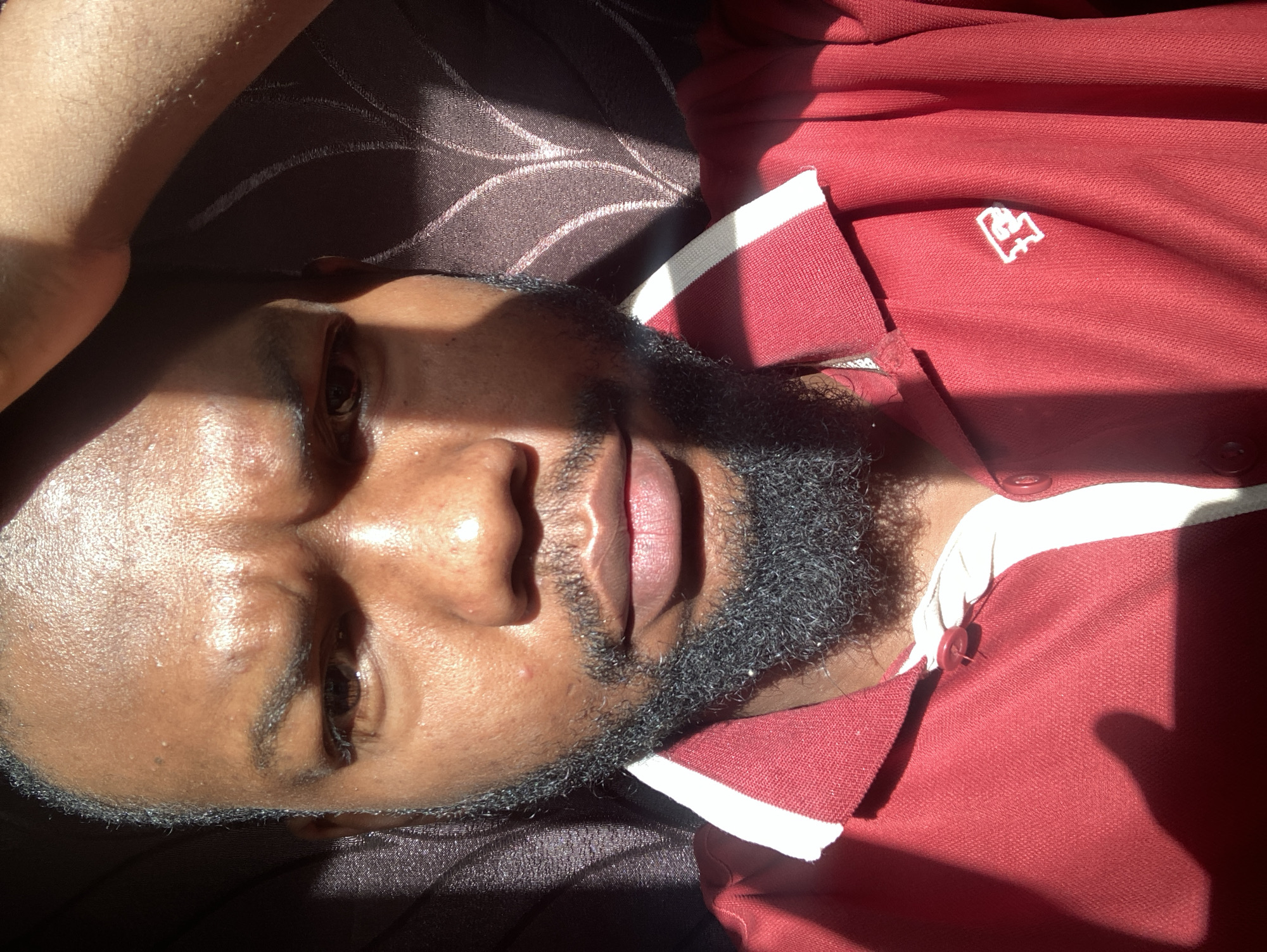 A medium shot, taken from an elevated perspective, shows a person lying supine on a dark-colored fabric. The person has brown skin, a black beard, and is wearing a maroon collared shirt with white trim. The lighting is bright, casting shadows across the person's face. The environment appears to be indoors. There is no indication of the city of Jos, Nigeria.