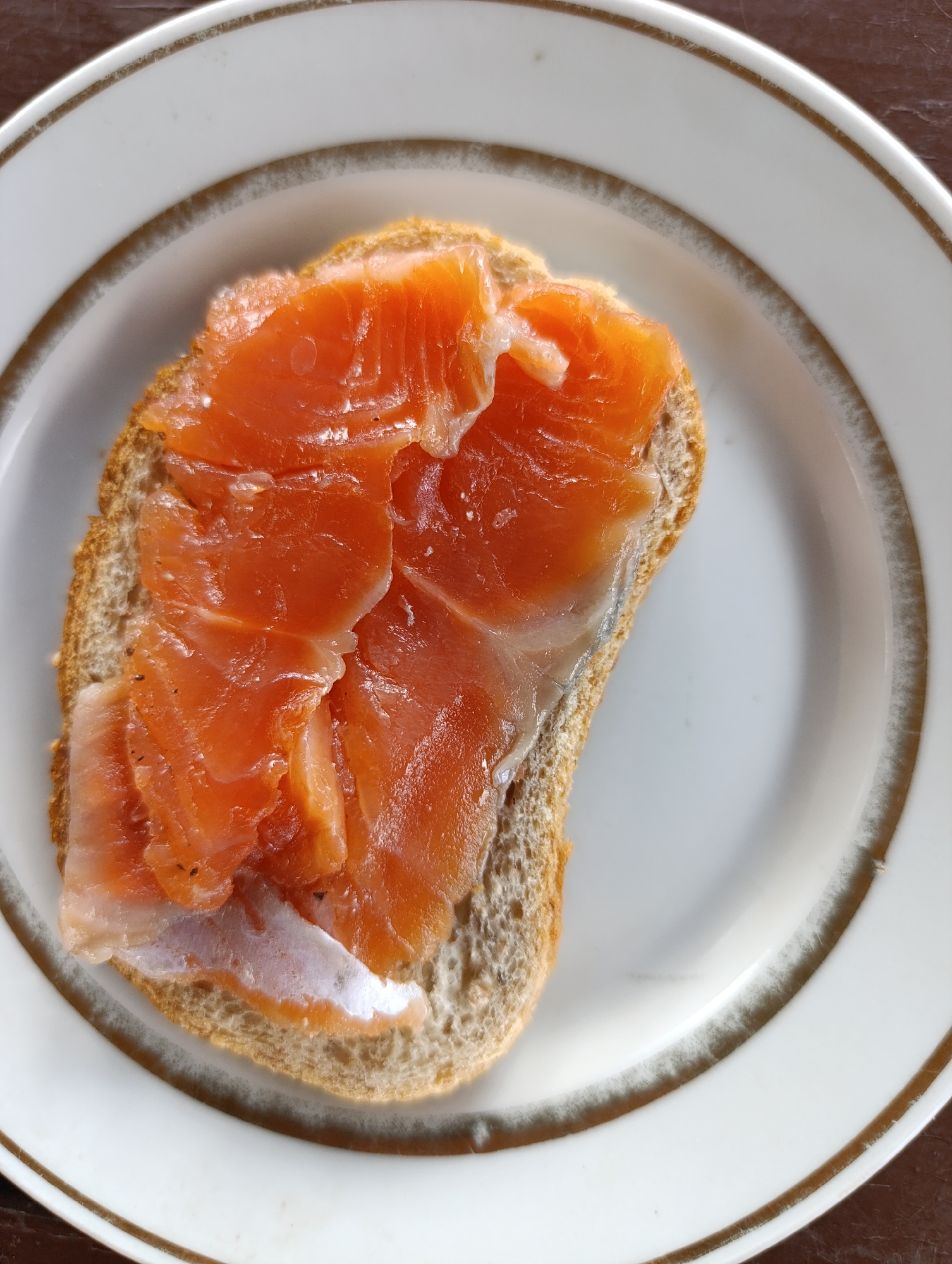 A close-up, overhead shot shows a slice of brown bread topped with thinly sliced raw salmon, placed on a white plate with a gold rim. The bread appears to be toasted and has visible texture from grains. The salmon is a bright orange color with streaks of white fat and some darker specks, possibly pepper. The plate has a visible concentric gold band around its edge. The background is a dark, out-of-focus surface, likely a table. The image was taken in Oktyabrsky SNT, Russia.