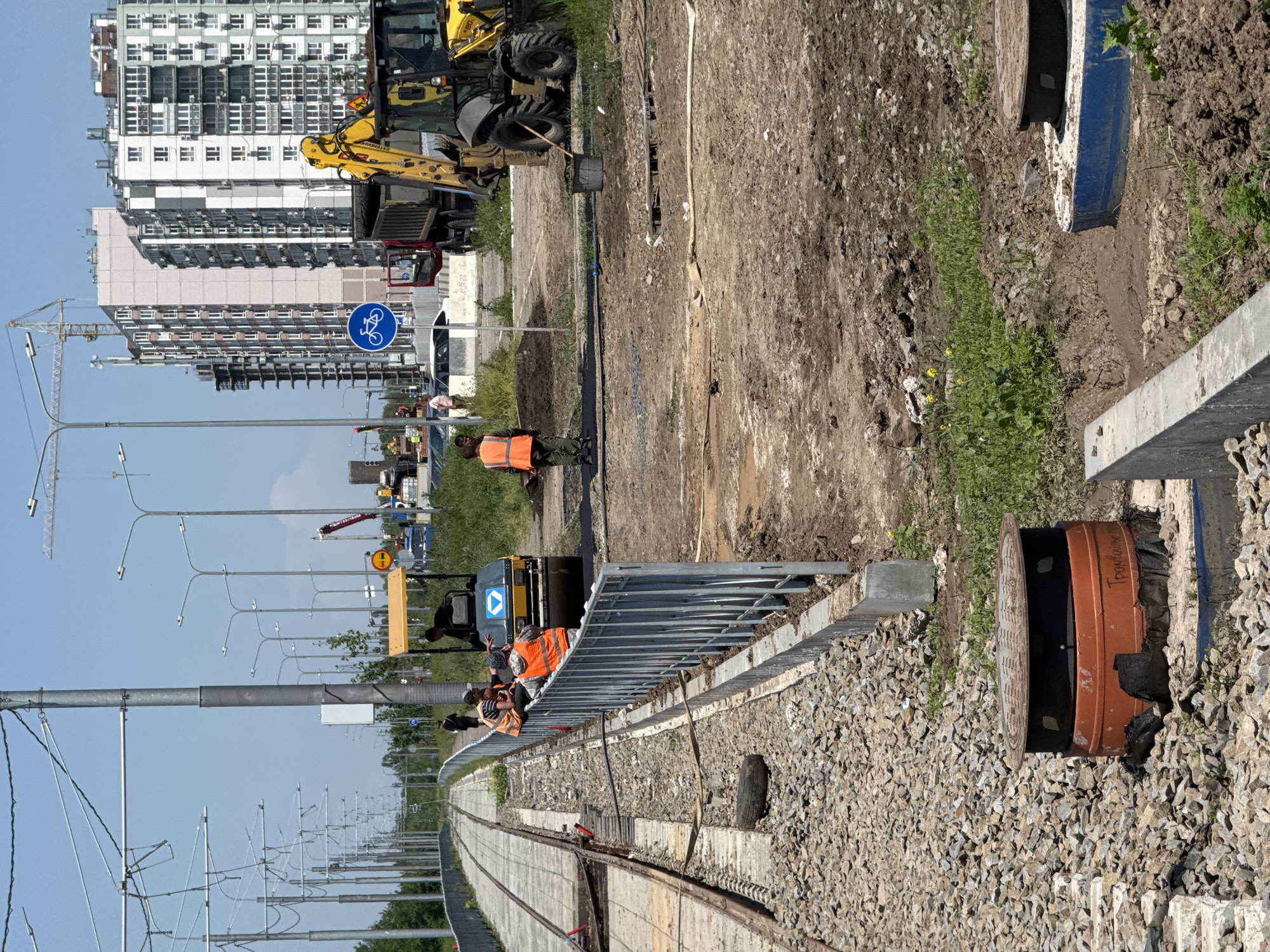 The image depicts a construction site in Rostov-on-Don, Russia. A railroad track is present, with workers in orange vests visible. Construction equipment, including a yellow backhoe and other machinery, are on-site. Buildings are in the background. Streetlights line the side of the road, and a bicycle sign is visible. A manhole cover is also visible in the foreground.