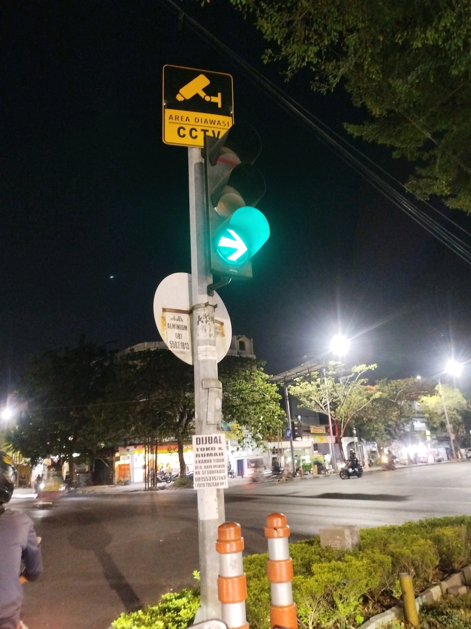 The image depicts a nighttime street scene in Surabaya, Indonesia, focusing on a traffic light pole. The traffic light is green with a right-pointing arrow illuminated. Attached to the pole is a yellow sign with black text and an icon indicating "AREA DIAWASI CCTV". Another circular sign below it has text that appears to be an advertisement for a property sale. The street is paved, and there are trees and buildings visible in the background, illuminated by streetlights. Several motorcycles and a blurry figure on a motorcycle are present on the road, suggesting moderate traffic. The overall environment is a typical urban street at night.