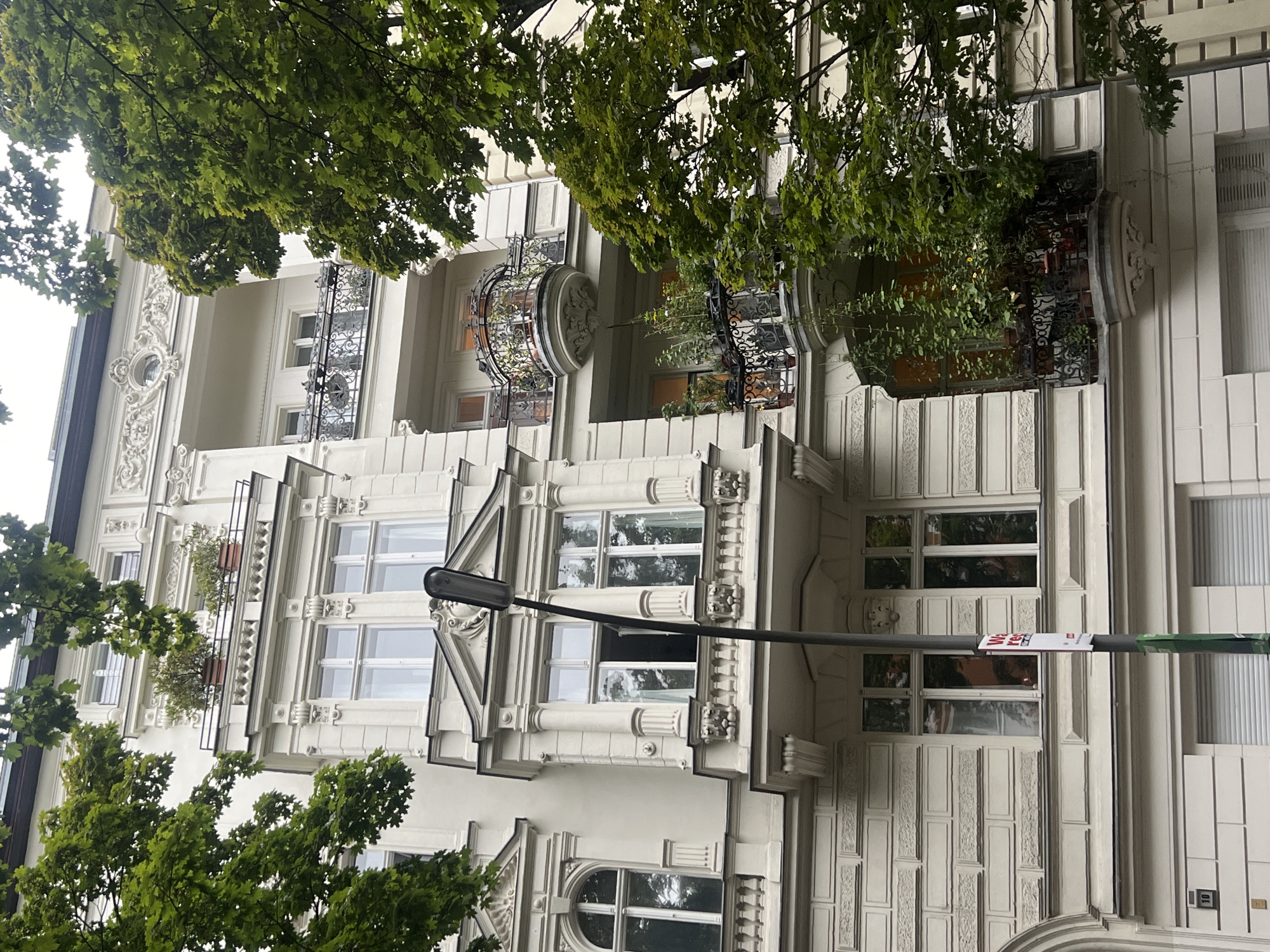 This image shows a light-colored building with ornate architectural details, common in Berlin, Germany. The building has multiple stories, each with balconies adorned with decorative railings. Some balconies are filled with potted plants and vines, giving them a lush, green appearance. The windows are large and appear to be multi-paned. A street lamp stands in front of the building, with a sign attached to its pole. Trees with green leaves are visible in the foreground and frame the building, suggesting an urban setting. The sky is overcast, indicating a cloudy day. The overall scene depicts a residential building in a European city, likely Berlin, with a touch of greenery and classic architectural style.