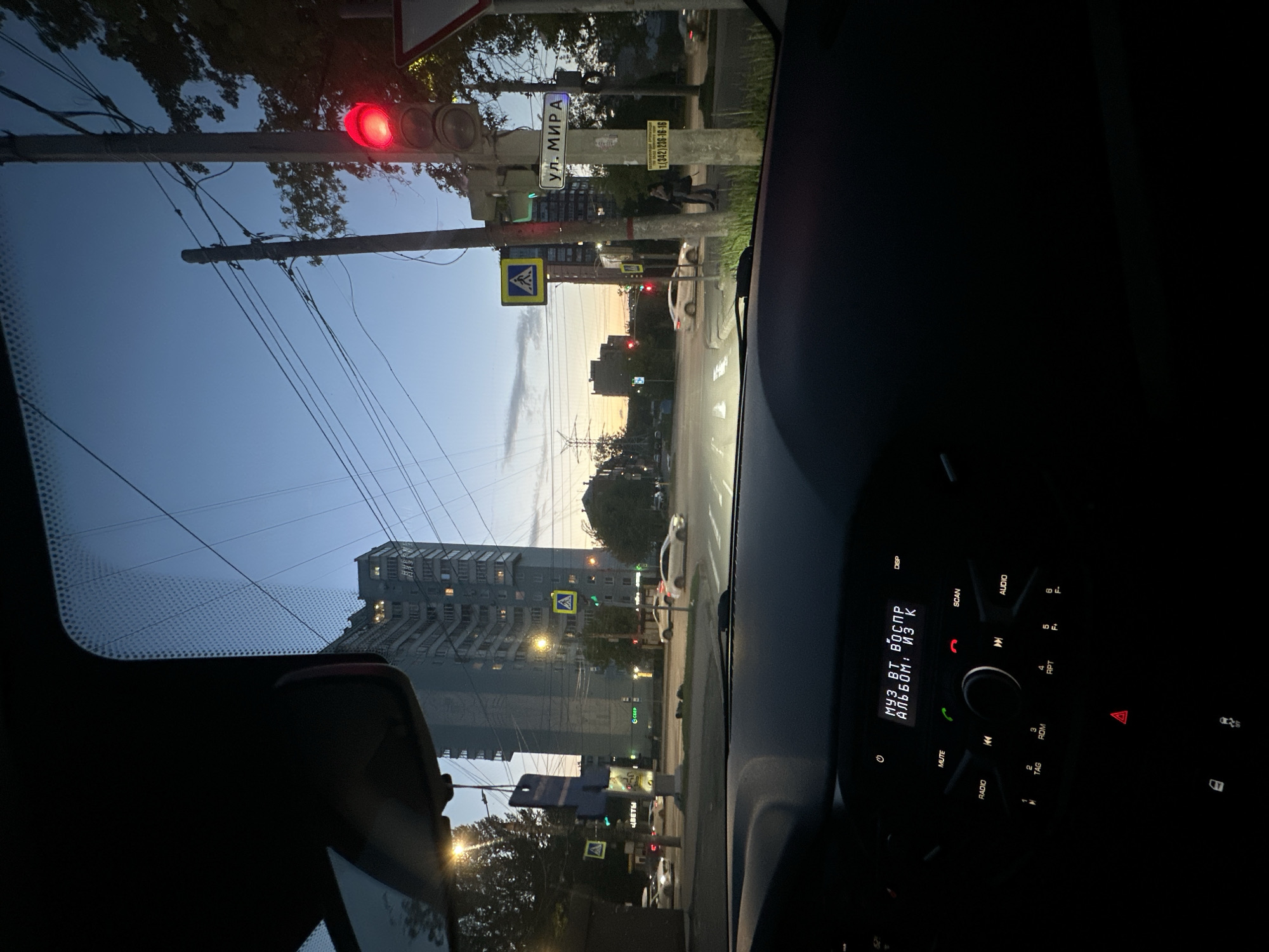 The view from inside a vehicle at dusk, likely in Perm, Russia, given the street sign reading "ул. МИРА" (Ulitsa Mira, or Peace Street). A red traffic light is visible ahead. Other signs and buildings are present. A pedestrian crossing sign is noticeable, indicating pedestrians might be nearby. The car's dashboard is partially visible with a radio display showing Cyrillic text.