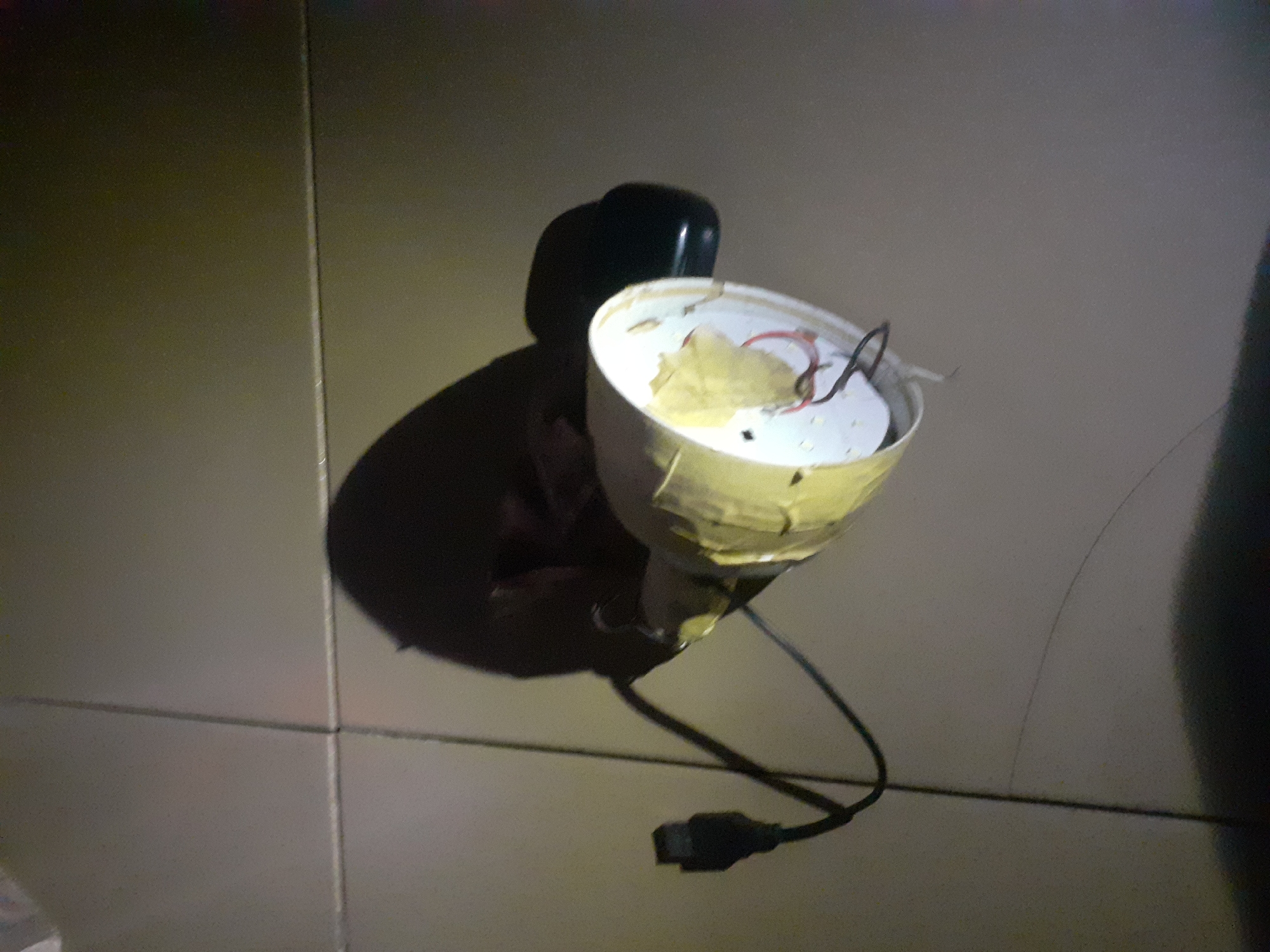 The image depicts a close-up, overhead view of a disassembled electronic device, likely a lamp or a projector, lying on a tiled floor. The device's casing appears to be made of white plastic, and it is partially covered with tape. Visible wires, some red and black, are connected to the internal components. A black power cord with a rectangular connector is also attached to the device. The background consists of light-colored, square tiles. The lighting in the image is uneven, with a bright light source illuminating the device from the side, creating shadows and highlighting the textures of the objects and the floor. There is no visible indication of the specific location within Jalingo, Nigeria; the scene appears to be indoors.
