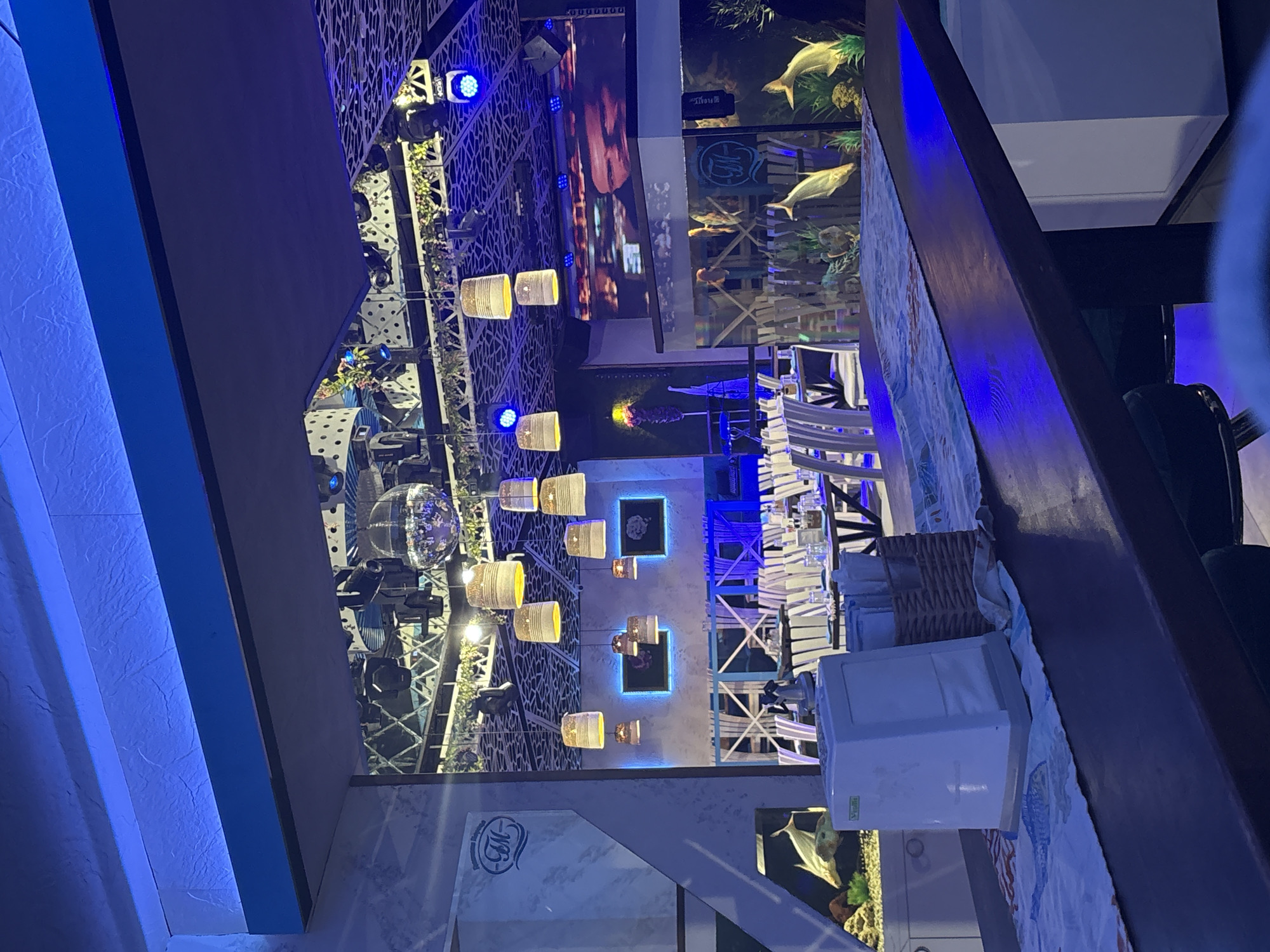 This image depicts the interior of a modern and atmospheric establishment, likely a restaurant, bar, or event venue, set in Plovdiv, Bulgaria, during the evening or night. The space is dominated by striking artificial lighting, featuring deep blue LED strips along architectural elements and spotlights, complemented by numerous suspended cylindrical light fixtures emitting a warm amber glow.
The decor includes intricate, geometrically patterned panels on the ceiling or upper walls. A large disco ball is visible on the left, suggesting a lively or entertainment-focused environment. Towards the right, a dining area is set with white tables and chairs, which feature slatted backs and some appear to have blue seat cushions. A woven basket and a white napkin dispenser are present on one of the tables.
A prominent feature of the venue is the integration of large aquariums, housing fish (possibly large freshwater species) and artificial aquatic plants, adding a unique, serene element to the modern design. The word "FLOAT" is visible on one of the aquarium structures. A large display screen or television panel is mounted on a wall, showing some content, while smaller, framed pictures with blue backlighting also adorn the walls. A stylized logo, possibly "MIK," is visible on a marble-like surface. No people are present in the scene, which emphasizes the architectural and decorative elements of the venue.