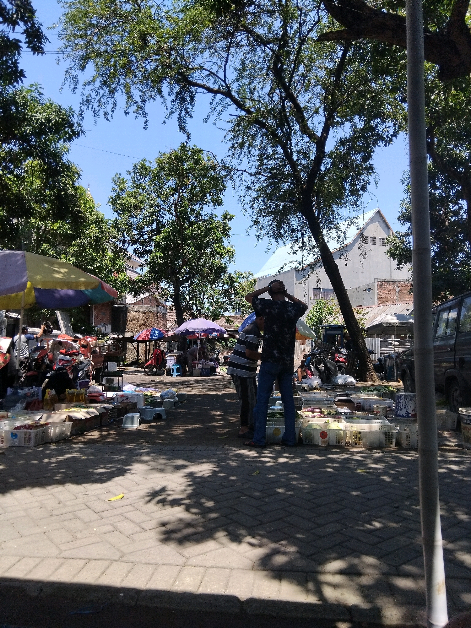 In Kepuh Kiriman, Indonesia, an outdoor market scene unfolds under a bright sky. The foreground is dominated by a paved walkway, dappled with the shadows of trees. White plastic crates, filled with produce such as eggplants and leafy vegetables, are displayed for sale. Several individuals are present. In the middle ground, two men stand near a collection of motorcycles and market stalls shaded by colorful umbrellas. One man, wearing a dark, patterned shirt and blue jeans, has his hands on the back of his head. Another man, in a striped shirt and dark pants, stands beside him. Further back, more stalls and a glimpse of a building with a light-colored roof and brick accents are visible. A dark-colored van is parked on the right side of the frame. The overall atmosphere suggests a bustling local marketplace.