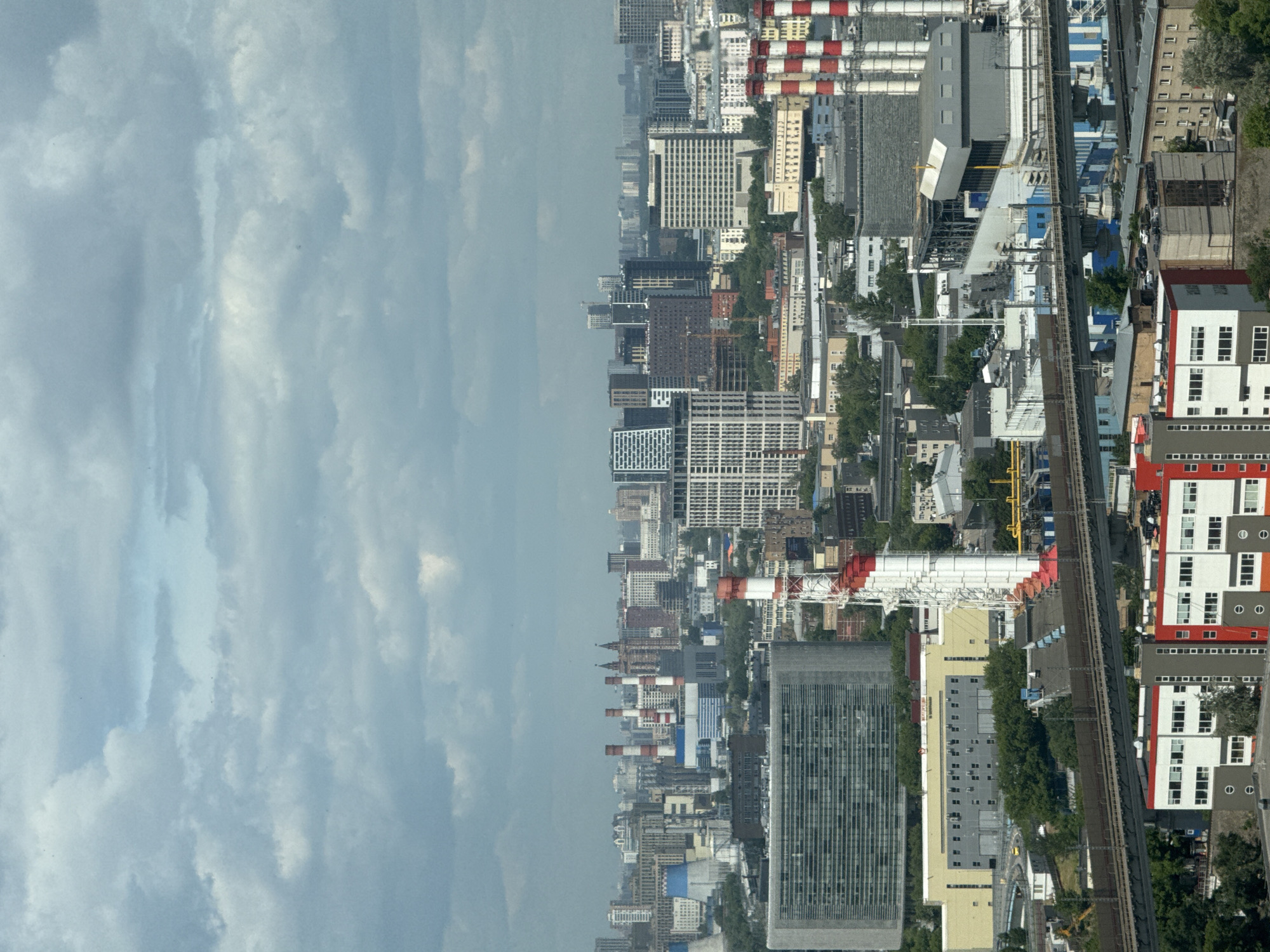 This image provides an aerial or high-angle view of an urban and industrial landscape, consistent with Khimki, Russia, captured during daytime under a bright, partially cloudy sky. The upper half of the frame is dominated by a light blue sky, filled with a patchwork of white and light gray clouds.
Below the horizon, a densely packed cityscape unfolds, featuring a diverse array of buildings. There are modern, tall high-rise structures with grid-like window patterns, alongside numerous multi-story residential and commercial buildings. A significant industrial presence is evident, characterized by large facilities and prominent red and white striped chimneys or pipes. Several yellow construction cranes are visible across the scene, indicating ongoing development.
A network of railway tracks, some appearing elevated, traverses the urban environment, weaving between buildings and industrial areas. Pockets of green foliage and trees are interspersed among the dense structures and along the railway lines. No specific human activity or readable text is discernible due to the distant, high-angle perspective.