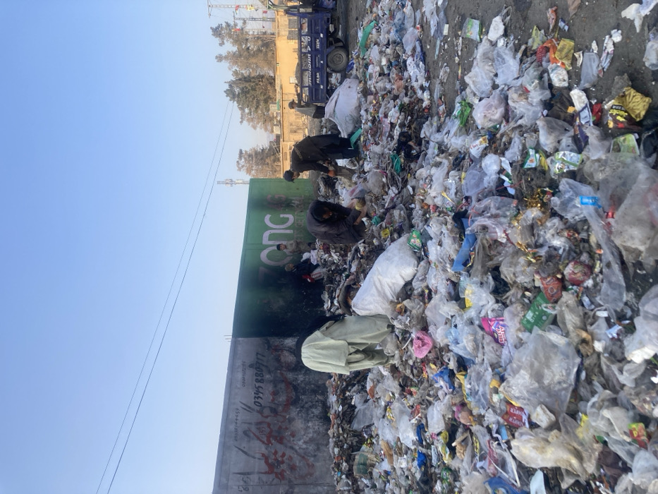 An outdoor scene in Quetta City Tehsil, Pakistan, depicts a large, unsorted accumulation of municipal solid waste occupying the foreground and midground. At least six male individuals are present within and around the refuse pile. Multiple individuals are actively engaged in sifting through the debris, bending or squatting, with at least two white sacks, presumably containing collected materials, visible on the ground. One individual in a light-colored shalwar kameez stands facing away from the camera near the center of the pile.
The background features a concrete boundary wall approximately three meters in height. The left section of the wall is gray, marked with black and red graffiti including Urdu script and the numeric sequence "0345 880 777." The right section of the wall is painted green and displays the white "ZONG" logo with "TELECOM" text below it. Two individuals are seated or squatting near the base of the green wall section.
To the far right, a light blue utility pickup truck, bearing the text "JHANDAR 2.0" on its side, is parked adjacent to the refuse pile. In the distant background, beyond the wall, sparse trees, utility poles, and power lines are visible against a clear, pale blue sky. The scene is illuminated by natural daylight.