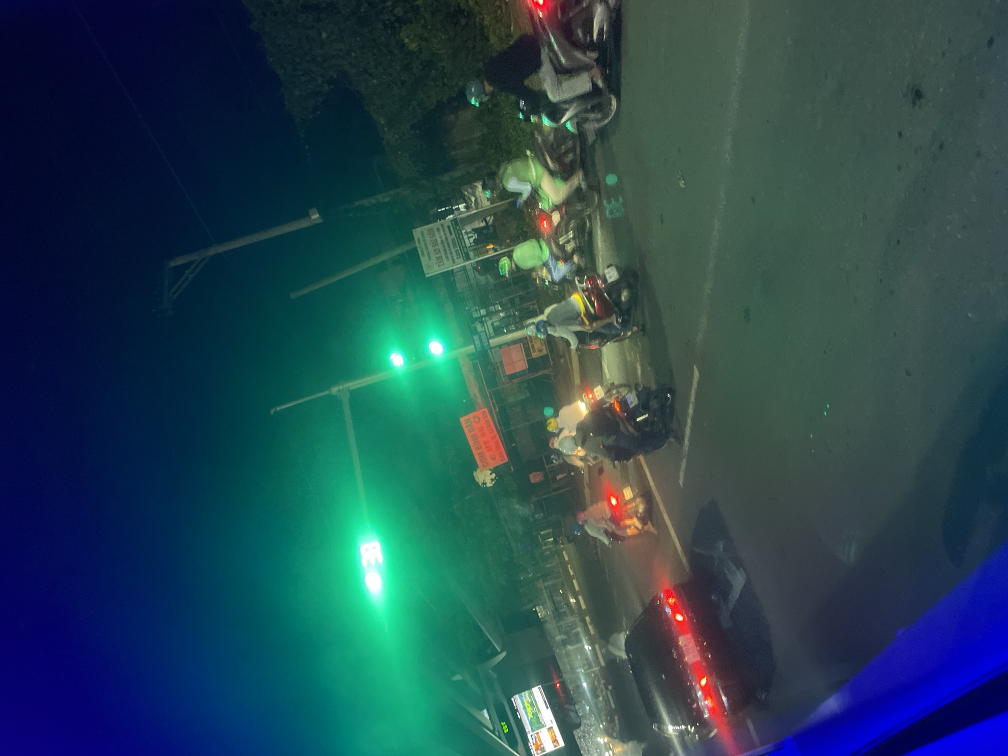 The image depicts a nighttime scene on a street in Binh Thuan Ward, Vietnam. A traffic light displays green, illuminating the area with a green hue. Several people are riding motorcycles, some wearing helmets. A black car is visible in the foreground, with its brake lights illuminated. In the background, there are buildings, trees, and a sign with Vietnamese text. The street is paved and appears to be an urban environment. The overall impression is one of traffic and movement at night.