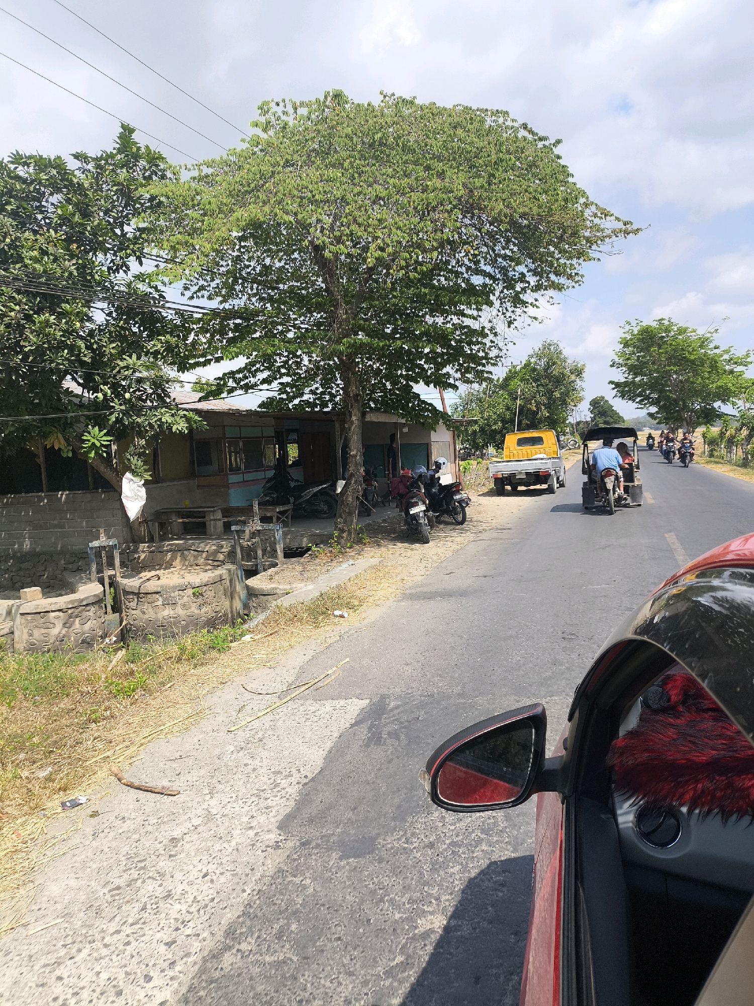 The image depicts a street scene in Sido Mulyo, Indonesia, on a sunny day. In the foreground, the interior of a red car is visible, with a red fuzzy accessory on the dashboard and the side mirror reflecting the road. The street itself is paved and asphalted, with dry grass and straw lining the side.
On the left side of the road, there is a building with a corrugated iron roof, painted in various colors including yellow and blue, with wooden window frames and a covered porch. Adjacent to the building are concrete structures, possibly part of a drainage system, with metal sluice gates. Several motorcycles are parked on the unpaved area in front of the building. A large tree with lush green foliage stands prominently near the building, casting a shadow on the road.
The road itself shows traffic. A yellow pickup truck is parked on the side, and further down the road, a three-wheeled vehicle, resembling a tuk-tuk, is carrying two passengers. Several other motorcycles with riders are also seen traveling on the road, moving away from the camera. The background consists of more trees and vegetation, with a partly cloudy sky overhead. The overall impression is of a rural or semi-urban area in Indonesia with typical local transportation.