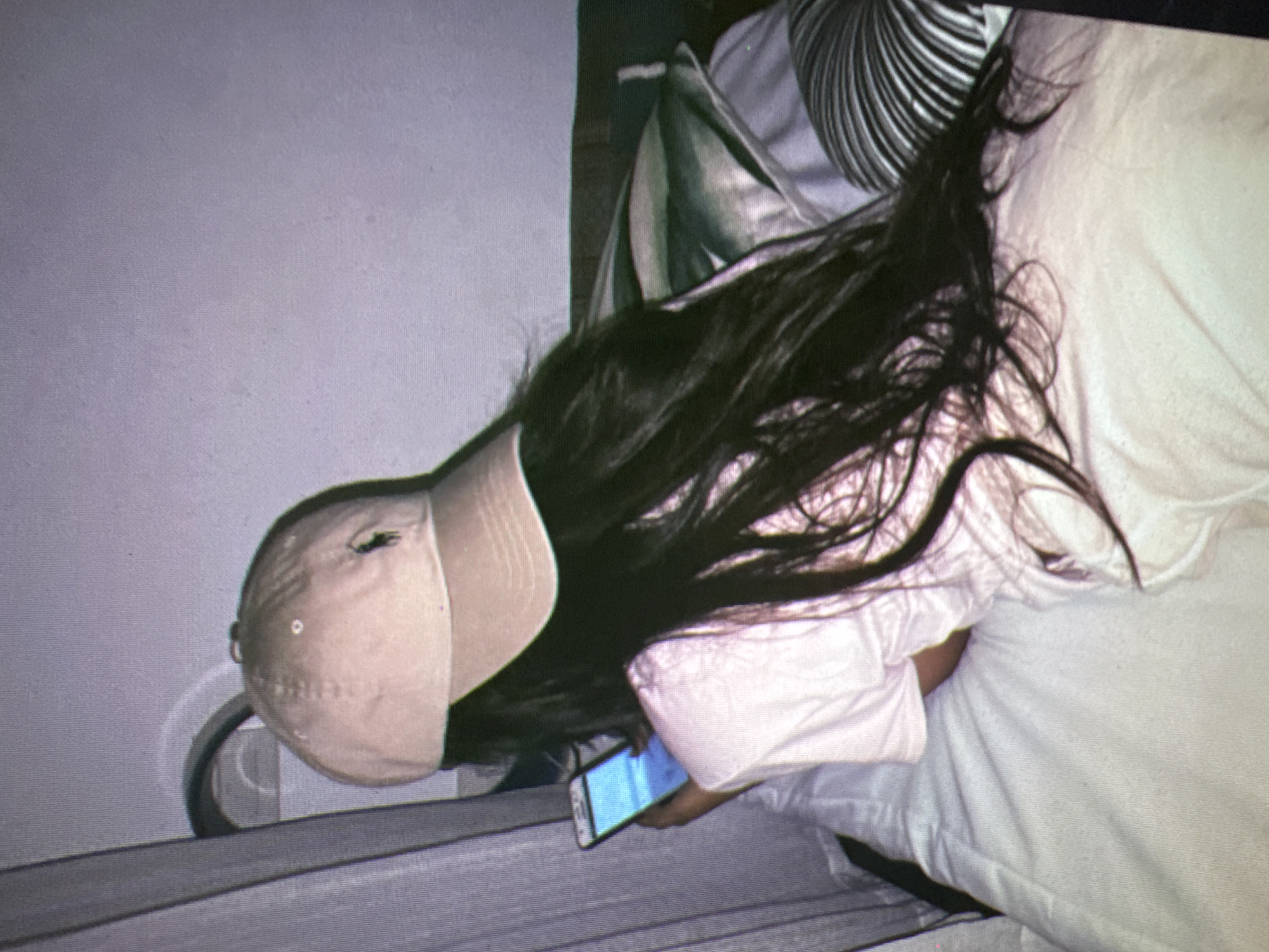 An overhead medium shot shows a person lying on a bed, looking at their phone. The person is wearing a light pink t-shirt and a beige baseball cap. They have long, dark hair that falls over their shoulders and down their back. The person's left arm is bent, and their hand is holding the phone, which is displaying a bright blue screen. The bed is covered in white linens. Behind the person, there are several pillows. One pillow in the background has a green and white striped pattern, and another pillow to the right has a black and white striped pattern. To the left of the person, a grey curtain is visible. The background wall is a plain, light color. The lighting in the scene is dim. The image is grainy and has a purple-grey tone. The location in the city of Castelo Branco, Portugal cannot be determined from the image.