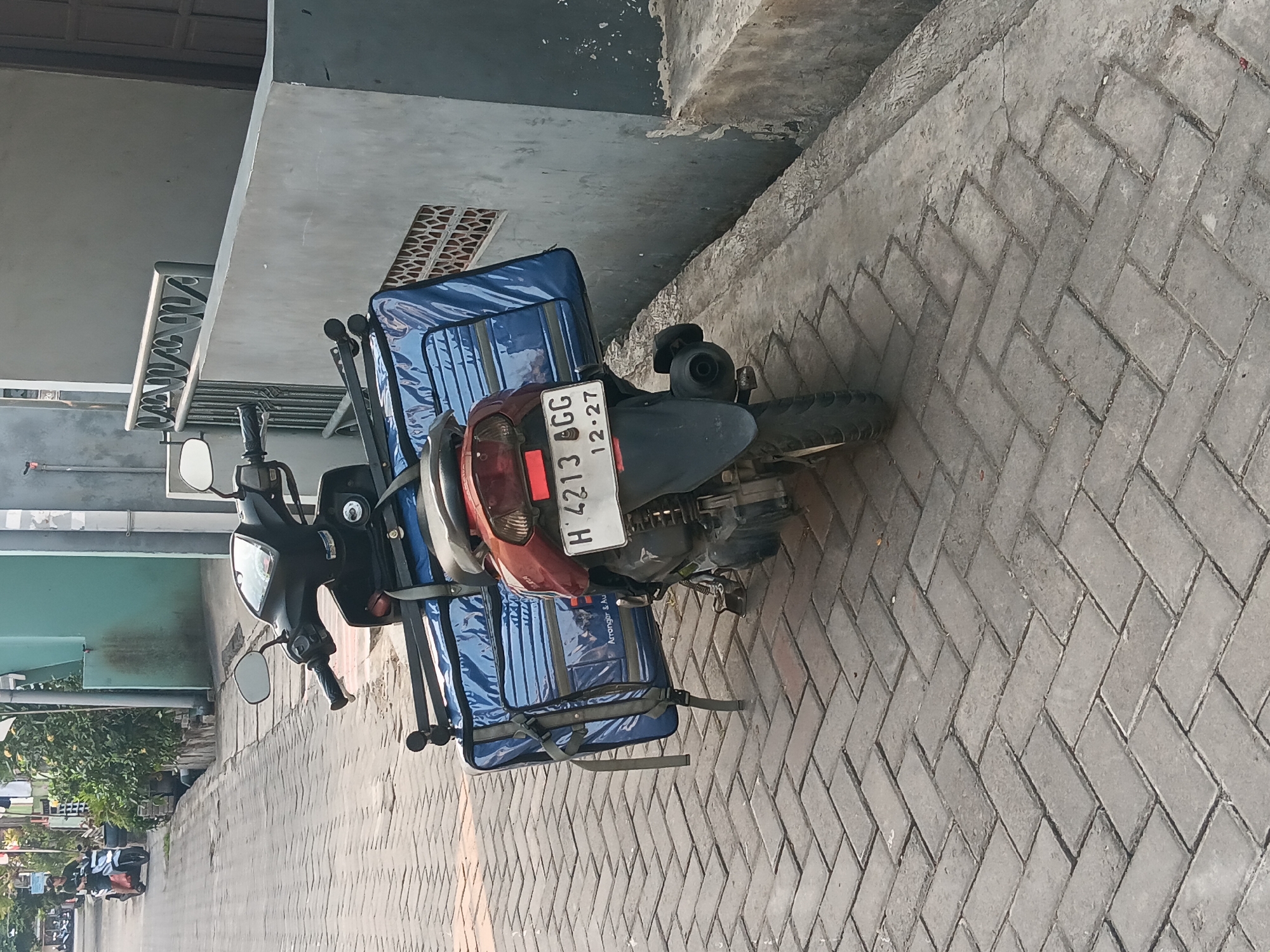 In Batursari, Indonesia, a parked red and black motorcycle is equipped with a large blue insulated delivery bag secured to its rear rack. The bag features a zipper closure and appears to be branded with "Arranger & Au". The motorcycle's license plate reads "H 4213 CGG 12.27". The vehicle is positioned on a patterned brick sidewalk adjacent to a concrete wall with a decorative metal railing. In the background, a narrow paved street with a drainage channel is visible, with another motorcycle and a person riding it in the distance. The environment suggests a residential or commercial street setting with some greenery.