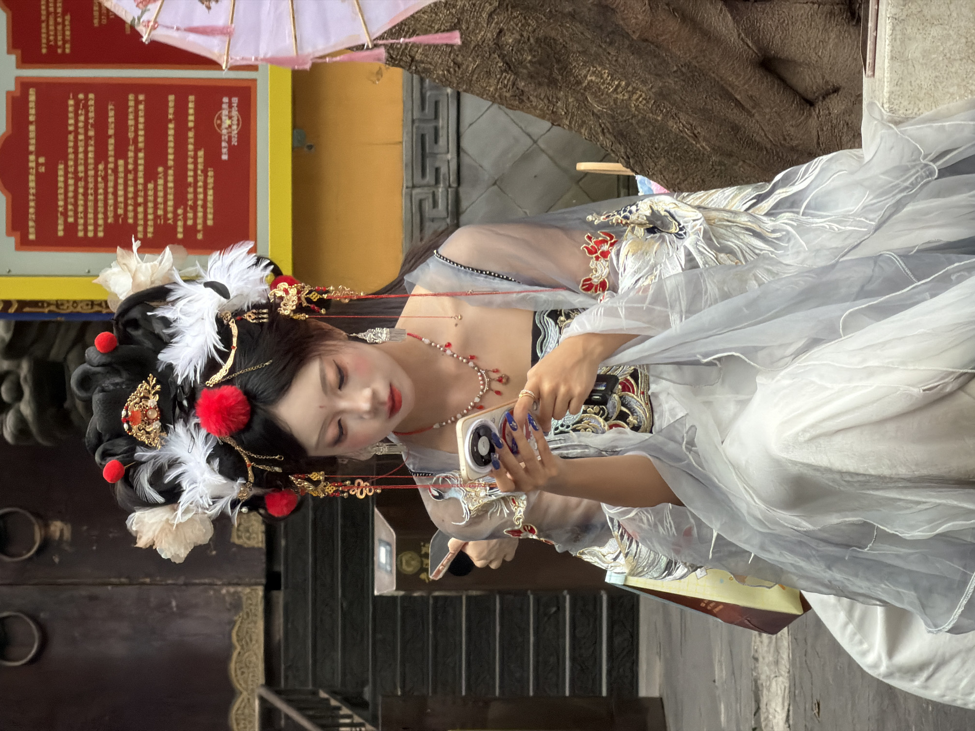 A young woman dressed in elaborate traditional Chinese attire, including a highly decorated headdress with feathers and red pom-poms, and a flowing grey and white gown with intricate embroidery, is seated outdoors and looking down at her smartphone. She is wearing heavy makeup, including dramatic eye makeup and red lipstick, and several pieces of jewelry. Her fingernails are painted blue with white accents. She is holding the phone in both hands, her thumbs poised to text or scroll. The background suggests an urban environment in Yuzhong District, China, with a traditional building facade visible on the left, including dark wooden doors with circular metal handles and stone steps. A yellow sign with red Chinese characters is on the right, partially obscured by the woman. A large, textured tree trunk is also visible on the right side of the frame. The overall impression is a juxtaposition of traditional culture and modern technology.