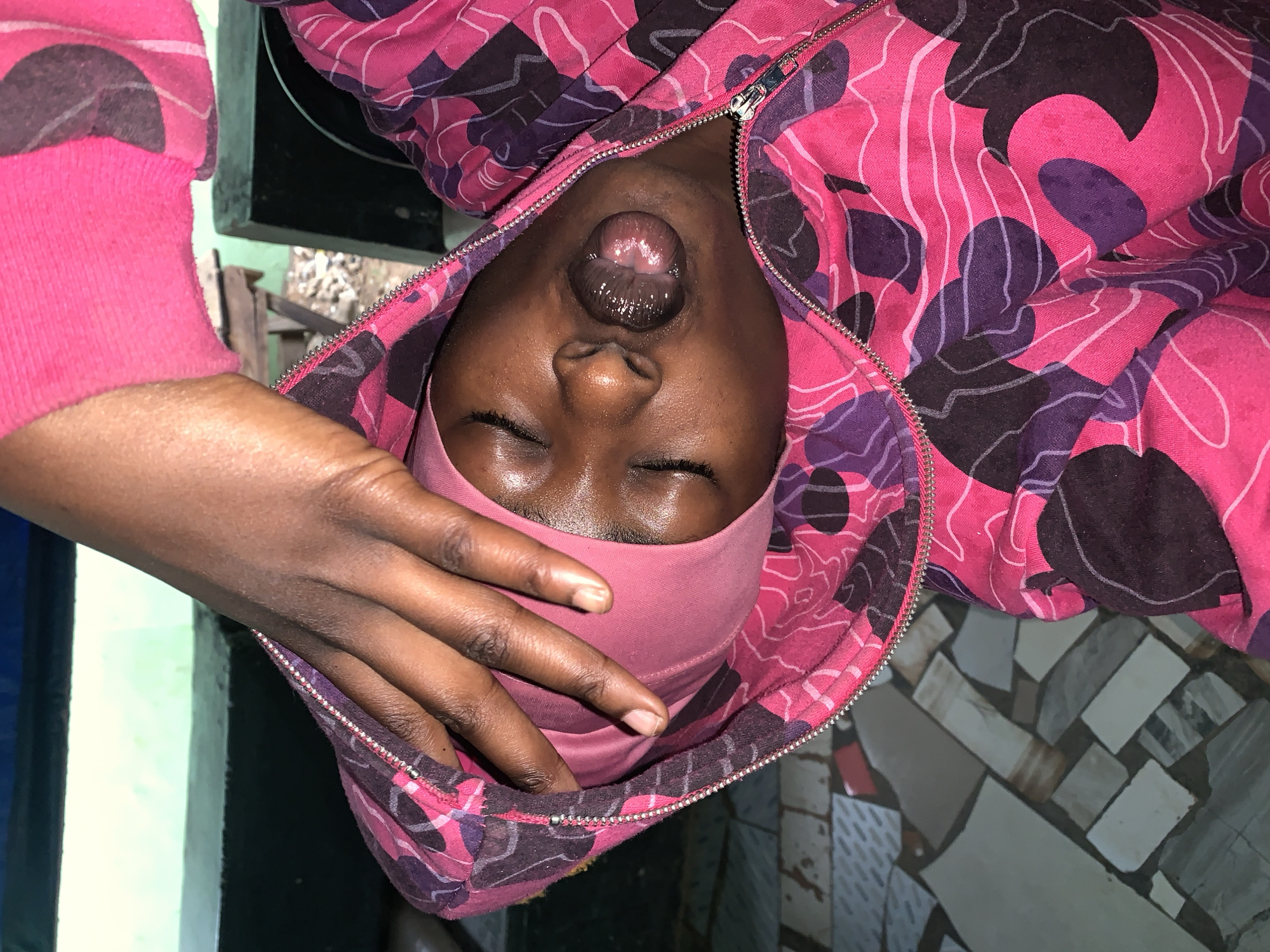 A person with dark skin is lying down wearing a pink head covering and a pink and purple patterned zip-up hoodie. The hoodie is partially unzipped, revealing the person's mouth. The person's right hand is near their face. The person is lying on a tiled surface. The image appears to be taken indoors, possibly in a residence near New Stadium, Nigeria. The location is inferred from the user's query about New Stadium, Nigeria.