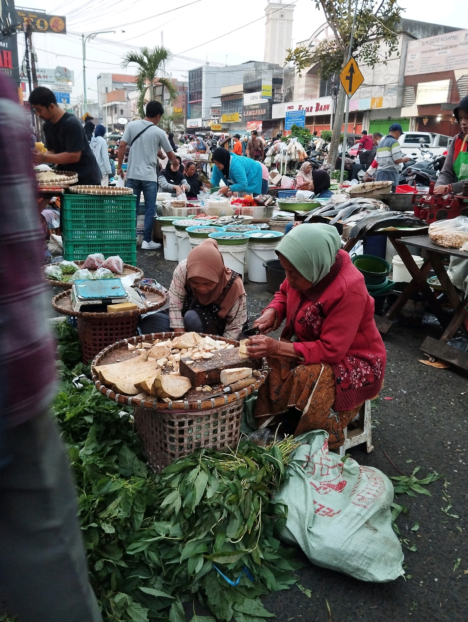The image depicts an outdoor market scene in Salatiga, Indonesia, likely a local produce market. In the foreground, two women are engaged in food preparation. One woman, wearing a brown hijab and apron, is seated on a low stool and appears to be slicing a large piece of white root vegetable or fruit on a wooden cutting board. She is using a knife and has several other similarly cut pieces on a woven basket in front of her. Beside her, another woman, also wearing a hijab, is seen from the side, potentially working on a similar task or arranging produce. In front of the women are woven baskets overflowing with fresh green leafy vegetables.
Behind them, various market stalls and activities are visible. There are stacks of green plastic crates, some filled with produce. Several men are also present, including one in a grey t-shirt and jeans who is walking away from the camera, and another in a black t-shirt who is attending to a stall with produce in woven baskets. In the background, there are rows of market stalls, buildings, motorcycles, and other people browsing or working. The ground is paved, and there are some scattered leaves and debris, indicative of an active marketplace. A large, light green fabric bag with red markings is placed on the ground near the produce. Traffic signs and overhead wires suggest an urban setting. The overall atmosphere is one of a busy, community-oriented market.