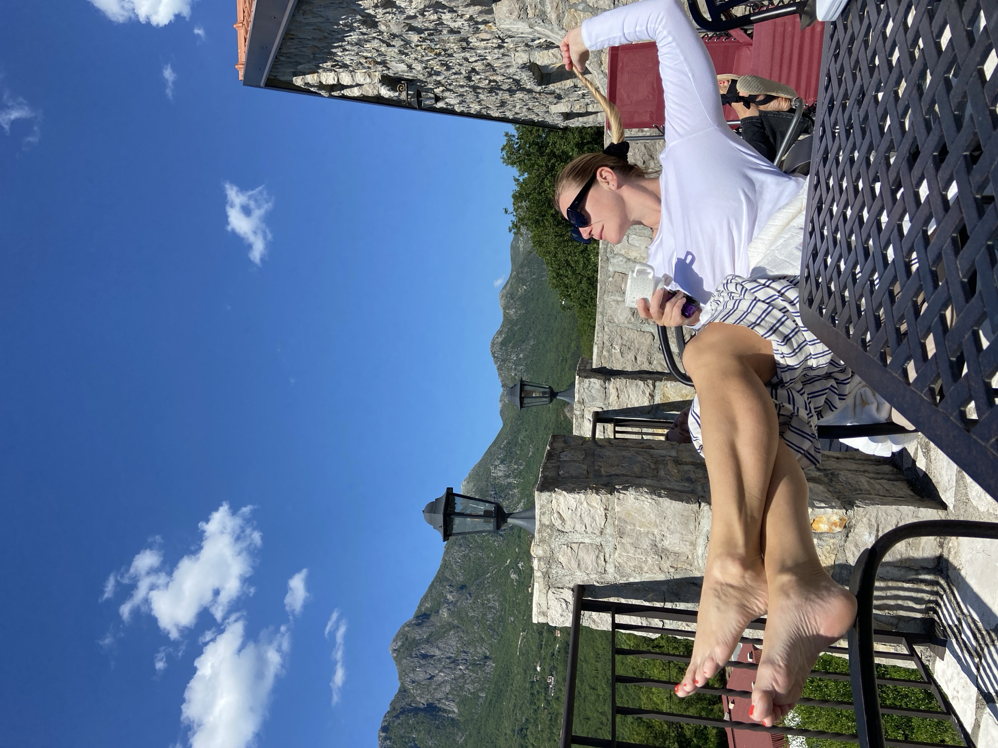 A woman sits at a black metal table on a stone patio in Sotonići, Montenegro. She wears a long-sleeved white shirt, sunglasses, and a striped skirt. She holds a white mug in one hand and touches her blonde ponytail with the other. Her bare feet are visible. Behind her is a stone building and a view of mountainous terrain dotted with trees and stone lanterns on the patio. The sky is blue with scattered white clouds.