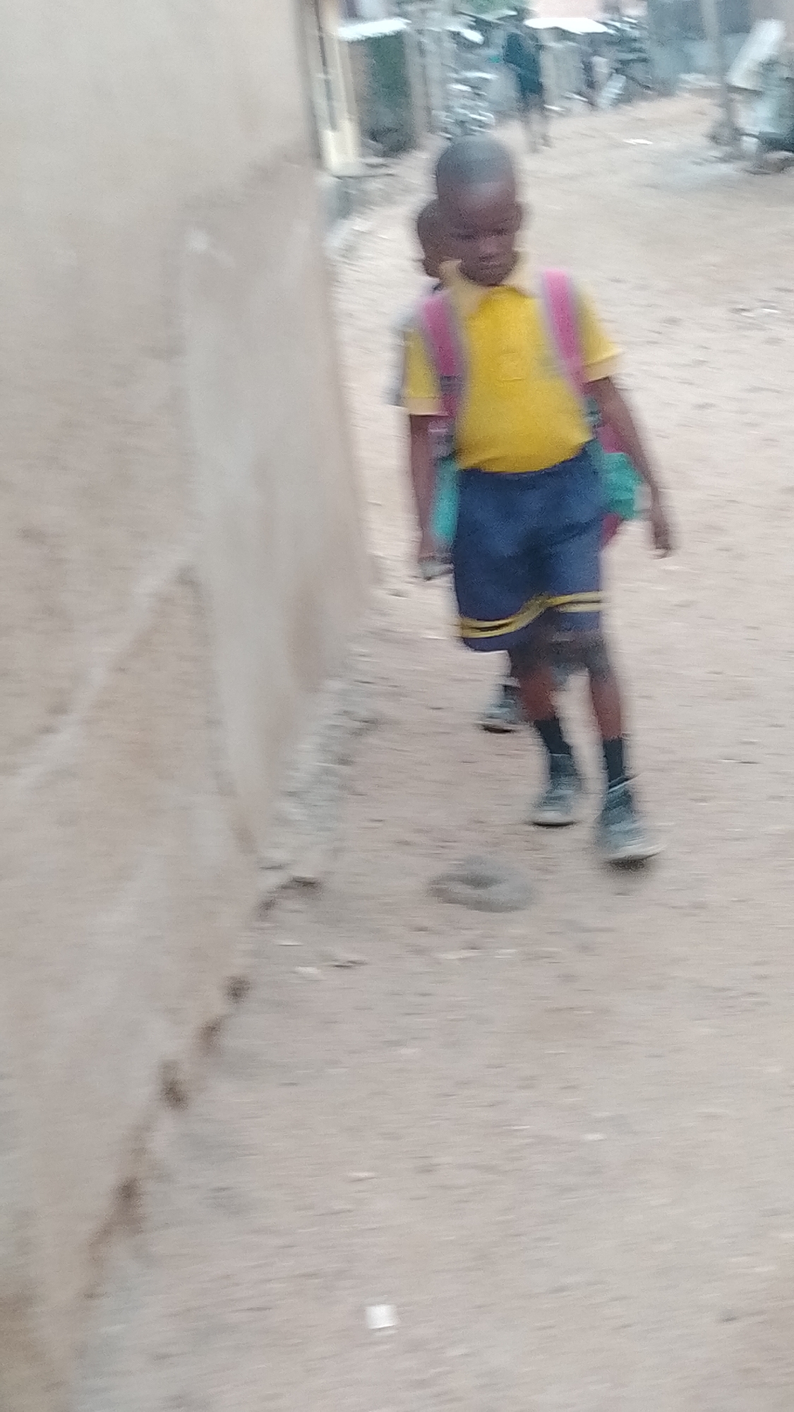 This image captures two young children, likely boys, walking along an unpaved path or street in Lokoja, Nigeria. The child in the foreground is more prominent, wearing a yellow polo shirt, dark blue shorts with yellow stripes at the hem, dark socks, and sneakers. He carries a pink backpack, with what appears to be a green item attached or held near his left side. A second child, wearing a similar yellow shirt, is partially visible behind the first, also appearing to be in uniform.
The children are in motion, suggested by the slight blur in the photograph, implying they are walking, possibly to or from school. The setting appears to be a local street, with a light-colored, textured wall (possibly mud or concrete) running along the left side of the frame. The ground is a dusty, uneven dirt surface. In the blurry background, other structures and buildings are discernible, indicating a residential or semi-urban environment. The lighting suggests it is daytime, but no strong shadows are present, hinting at an overcast sky or a time of day with muted sunlight. There is no visible text in the image.