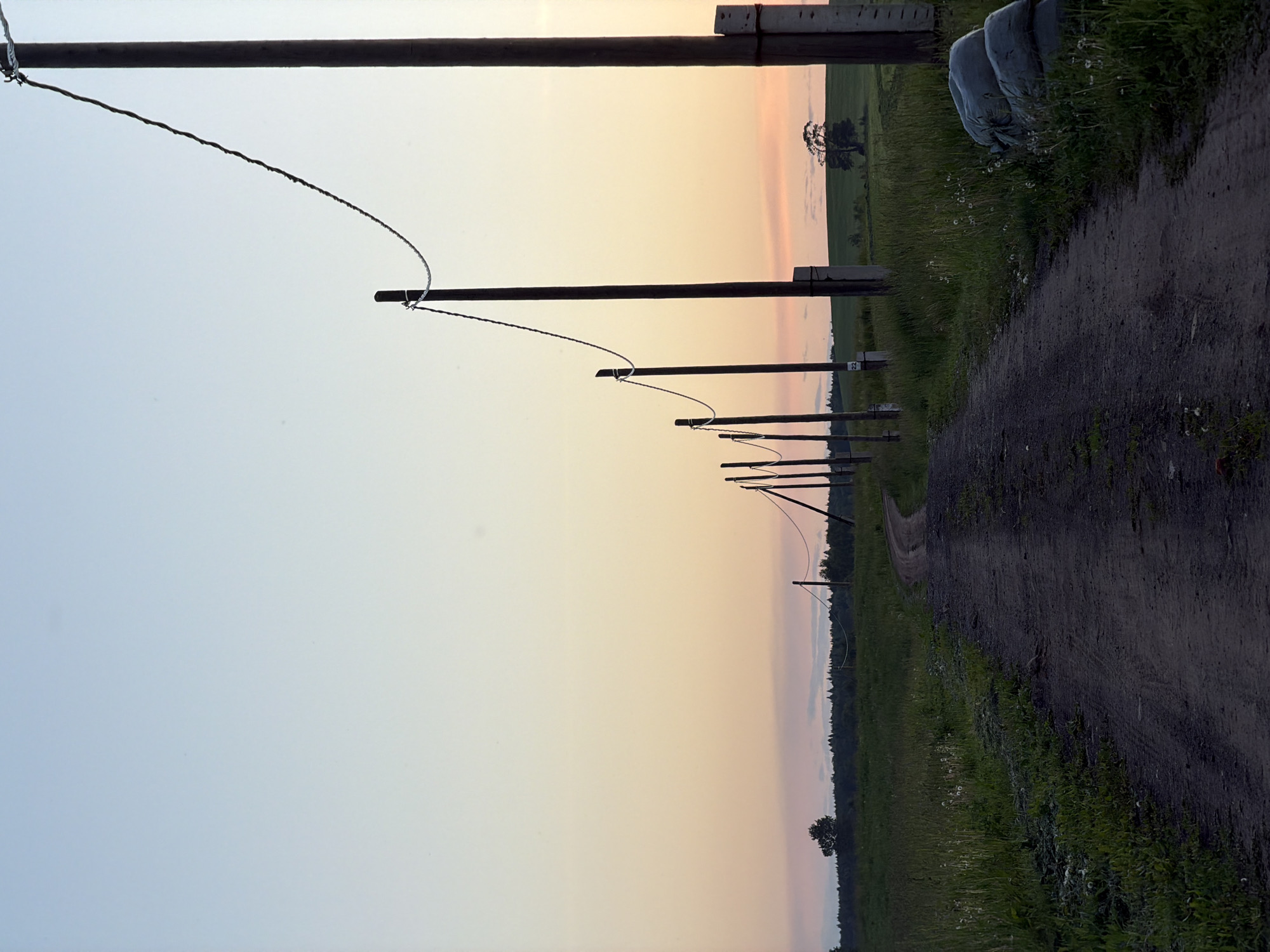 The image captures a rural landscape in Kechur, Russia, likely at dawn or dusk, characterized by the soft gradient of colors in the sky, ranging from light blue to a pale orange-pink. A dirt road occupies the right side of the frame, curving gently as it recedes into the distance. To the left of the road, a green grassy field stretches out.
Along the left edge of the scene, a series of wooden poles, possibly fence posts or rudimentary utility poles, stand in a line, with thin wires strung between them, creating a rhythmic pattern. In the far distance, a dark horizon line suggests a treeline or forest, and a single, isolated tree is visible in the field.
Near the foreground on the right, close to the dirt road and partially obscured by grass, are several light-colored sacks or bags. One of the wooden poles has the number "22" inscribed or attached to it. The overall atmosphere is quiet and serene, with no human activity visible. The weather appears clear.