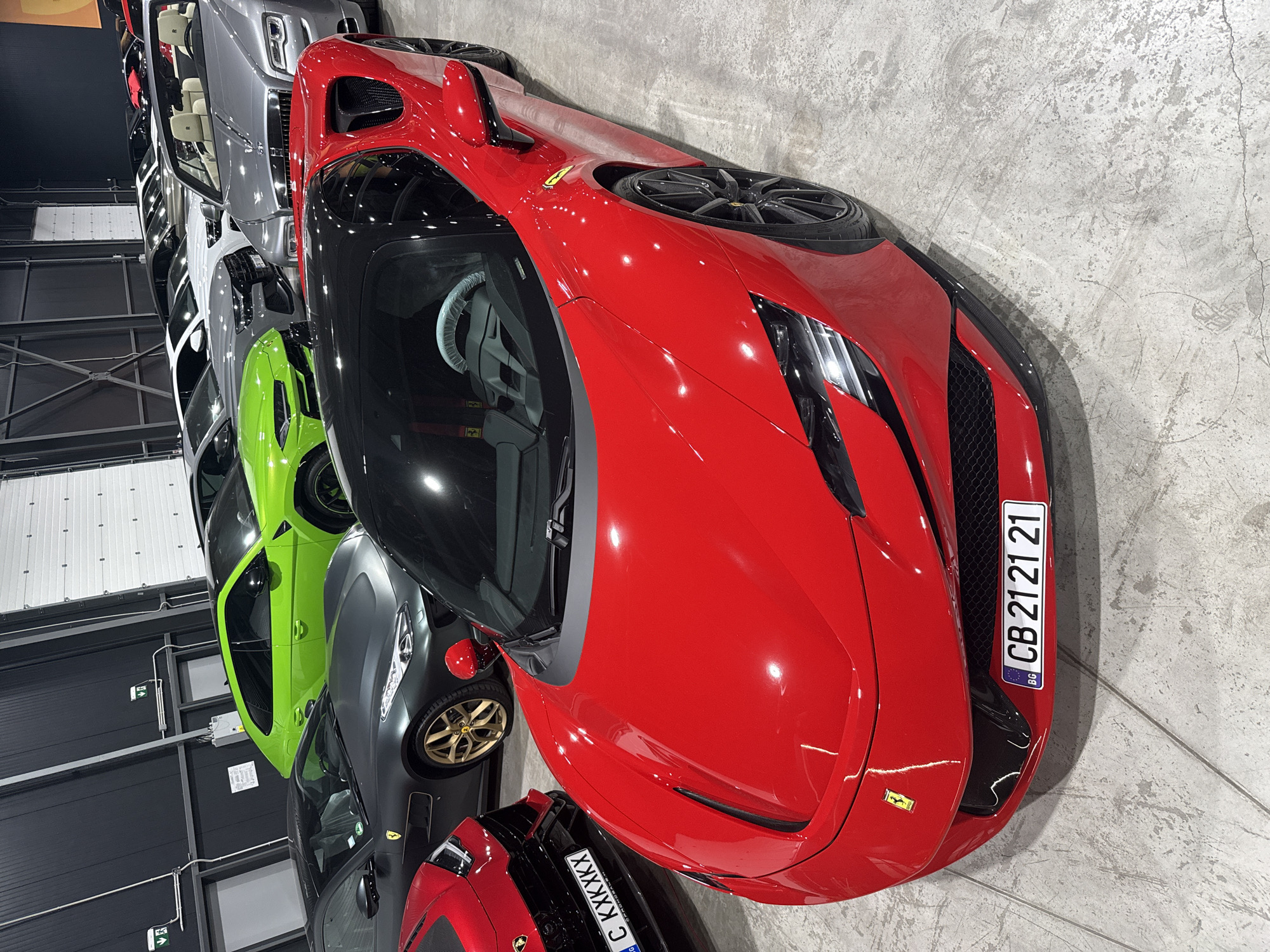 The image depicts a showroom or a private garage filled with various luxury vehicles. In the foreground, a bright red Ferrari sports car is prominently displayed, its sleek design and shiny paint reflecting the overhead lighting. Behind it, several other cars are visible, including a lime green Lamborghini SUV, a dark gray Ferrari coupe, and a silver Rolls-Royce convertible. Other vehicles, including a white Audi and a white Porsche, are parked further back in the rows. The environment appears to be indoors, with a concrete floor and a dark gray wall in the background, suggesting a commercial space or a high-end collection. The image is taken from a slightly elevated perspective, looking down at the cars. There are no people visible in the frame. The license plate of the red Ferrari reads "BG CB 21 21 21", indicating it is registered in Bulgaria. The location is not explicitly identified as Plovdiv, Bulgaria, but the license plate suggests it is within Bulgaria.