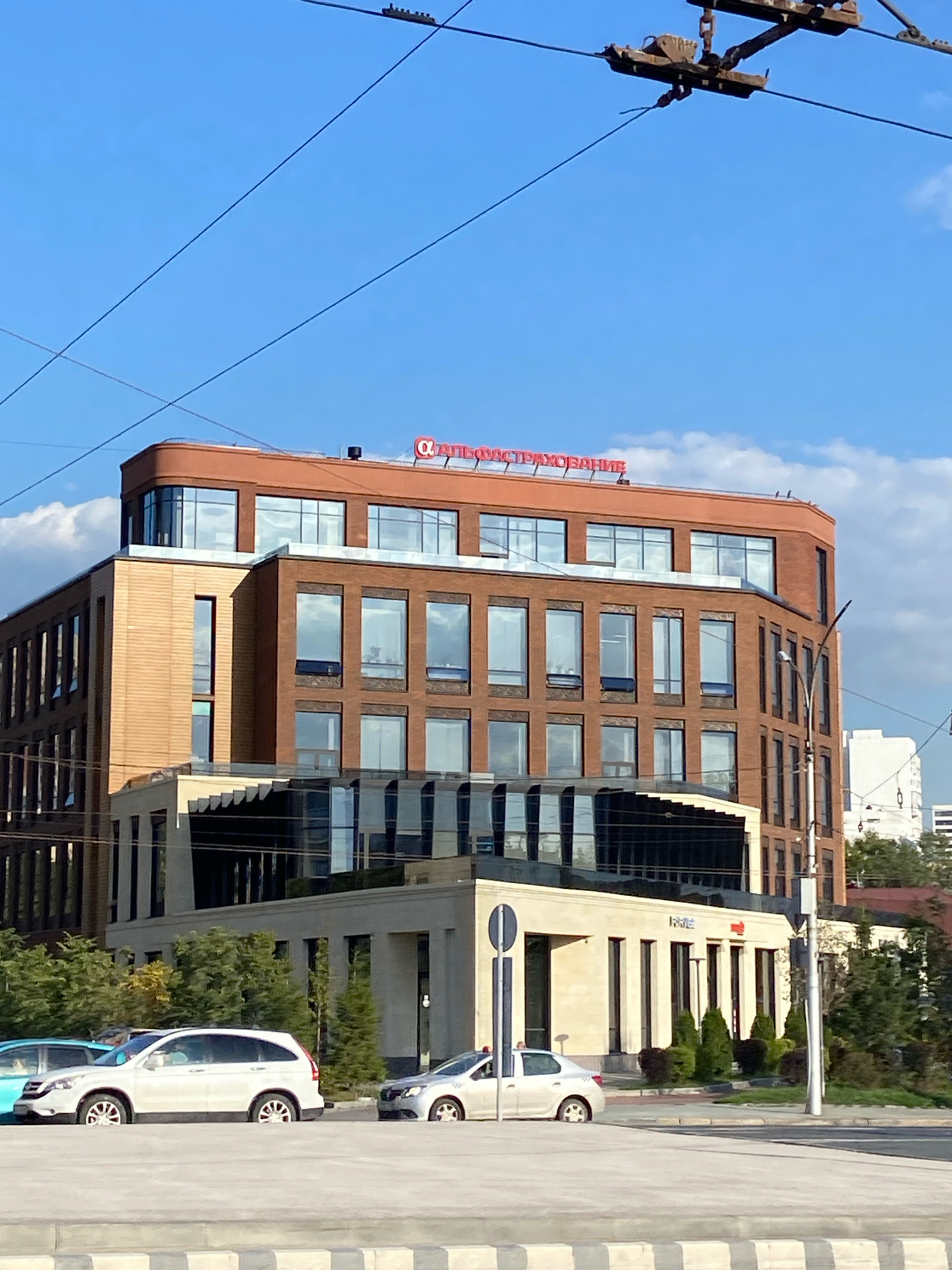 The image captures a modern, multi-story commercial building in Novosibirsk, Russia, with a distinctive red sign displaying Cyrillic text on its upper facade. The building features a combination of brickwork and lighter colored materials, with large, reflective windows that offer glimpses of the interior and the sky. In front of the building, there is a low-rise section with a more minimalist design, featuring vertical window openings. Several trees and bushes are visible, contributing to the urban landscaping. On the street in front of the building, two cars are parked: a white SUV and a silver sedan. The sky above is a clear blue with some scattered white clouds, and power lines are visible crossing the frame.
