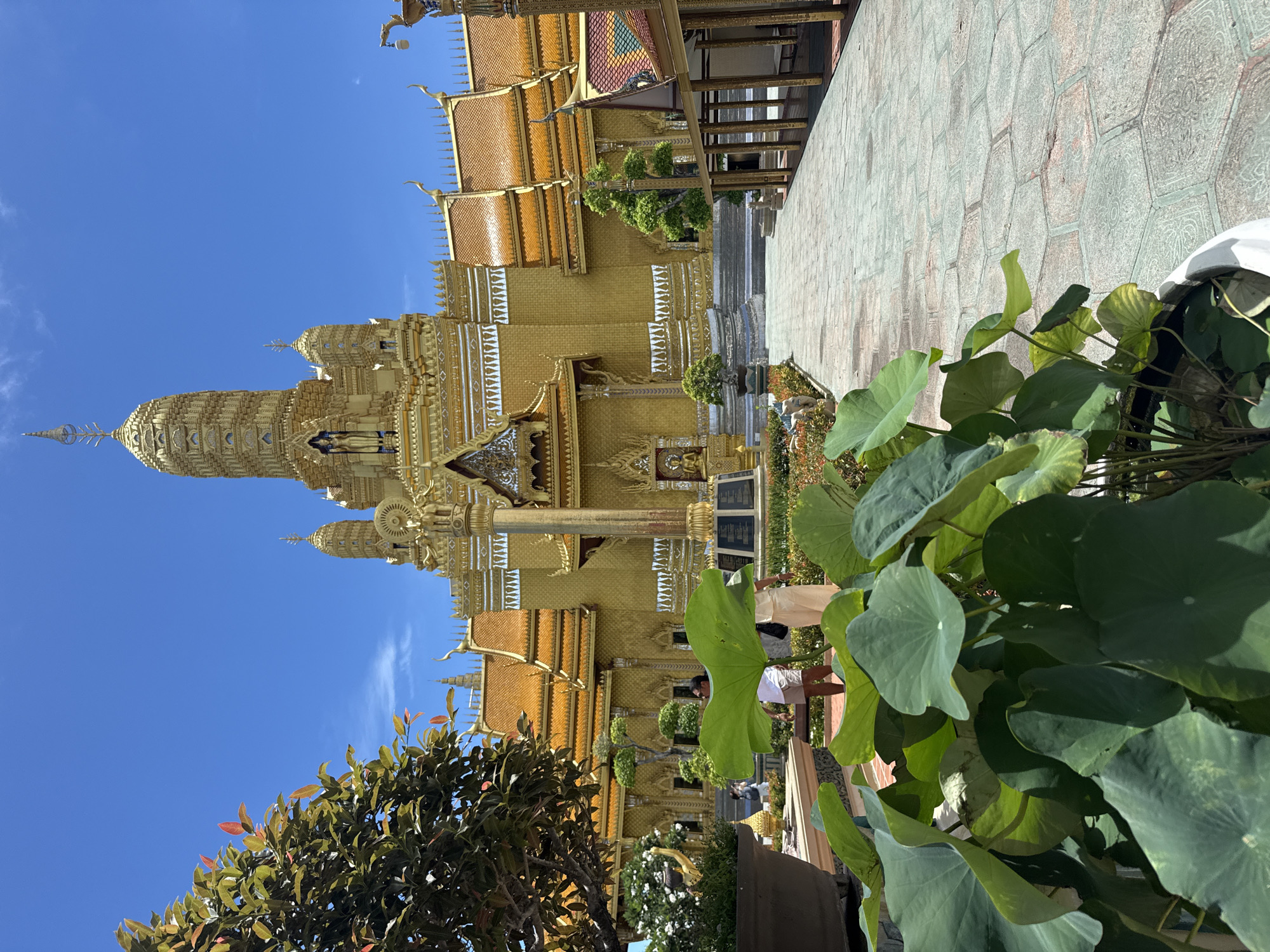 The image depicts a golden Thai temple under a clear blue sky, likely located in Bang Pu Subdistrict Municipality, Thailand. The temple is ornate and multi-tiered, featuring intricate details and a tall spire. A paved walkway is visible to the right of the temple, along with some foliage in the foreground, including large green leaves. Several people appear in the mid-ground near the temple.