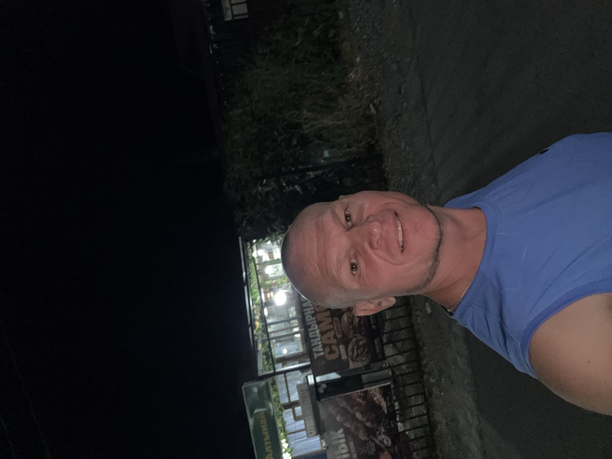 The image shows a man in a blue t-shirt standing outside at night. Behind him, there is a building with lights and signage. One sign is in Cyrillic script and appears to advertise food. There are also some plants and a fence. The lighting suggests it is evening or night. The location is identified as the city of Urdzhar, Kazakhstan.