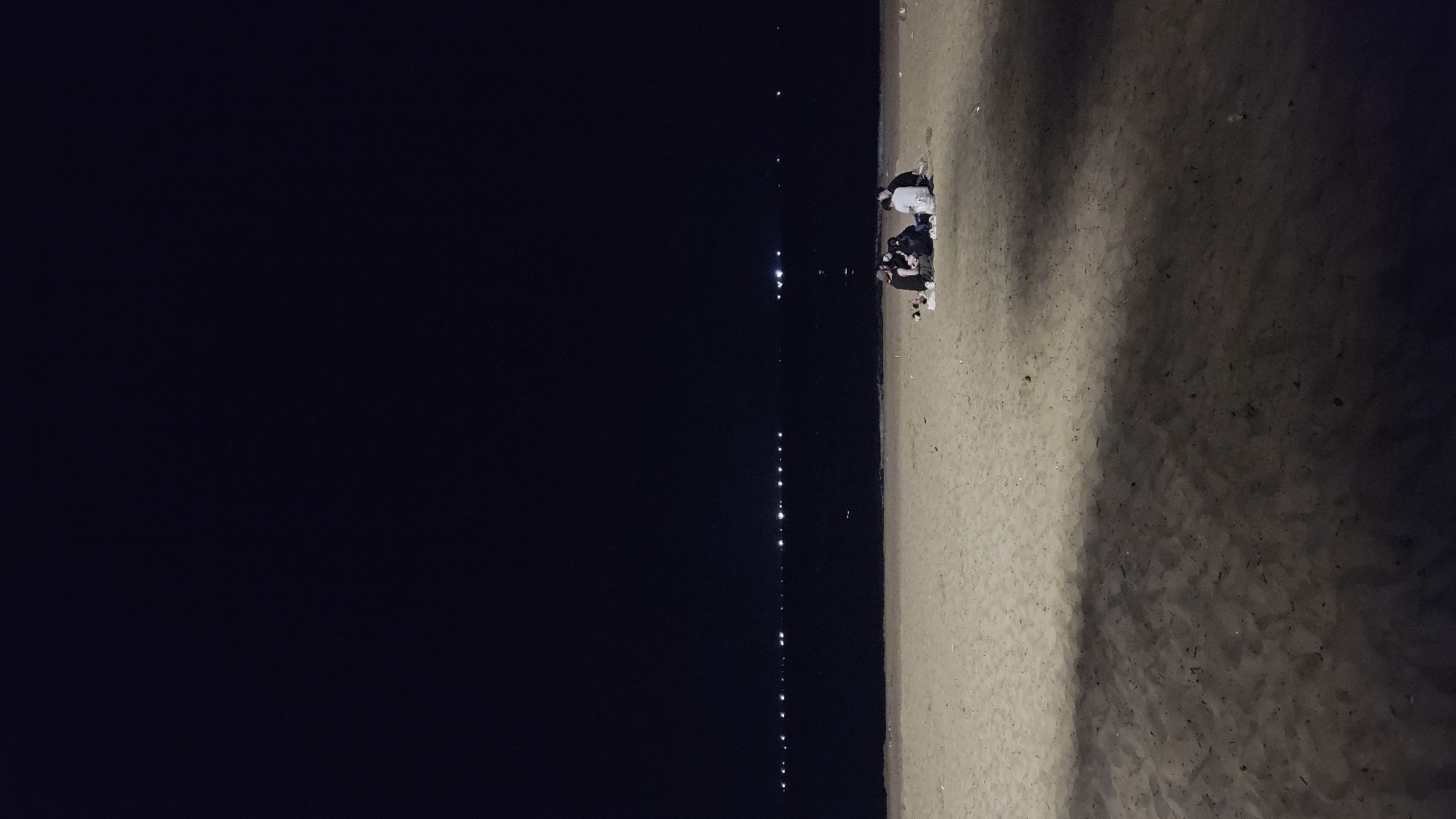 The image depicts a tranquil night scene on a beach, likely in Nha Trang, Vietnam. The frame is divided vertically, with a vast, dark body of water on the left and a sandy shore on the right.
On the dark water, a distinct vertical line of scattered, small bright lights shimmers, likely reflections from distant boats or coastal establishments, extending from the foreground into the background. The water itself appears calm, reflecting the sparse light sources.
On the sandy right side of the image, a small group of approximately three to four people is gathered near the water's edge. They are seated or kneeling on the sand, their figures somewhat indistinct due to the low light. One individual appears to be wearing a light-colored top, offering a slight contrast to the otherwise muted tones. The sandy texture of the beach is visible, with subtle variations in shade.
The activity appears to be a quiet gathering or relaxation by the sea at night. The individuals seem to be enjoying the peaceful ambiance, perhaps socializing or simply observing the dark expanse of the ocean and its distant lights. The overall atmosphere is calm and serene. There is no visible text in the image.