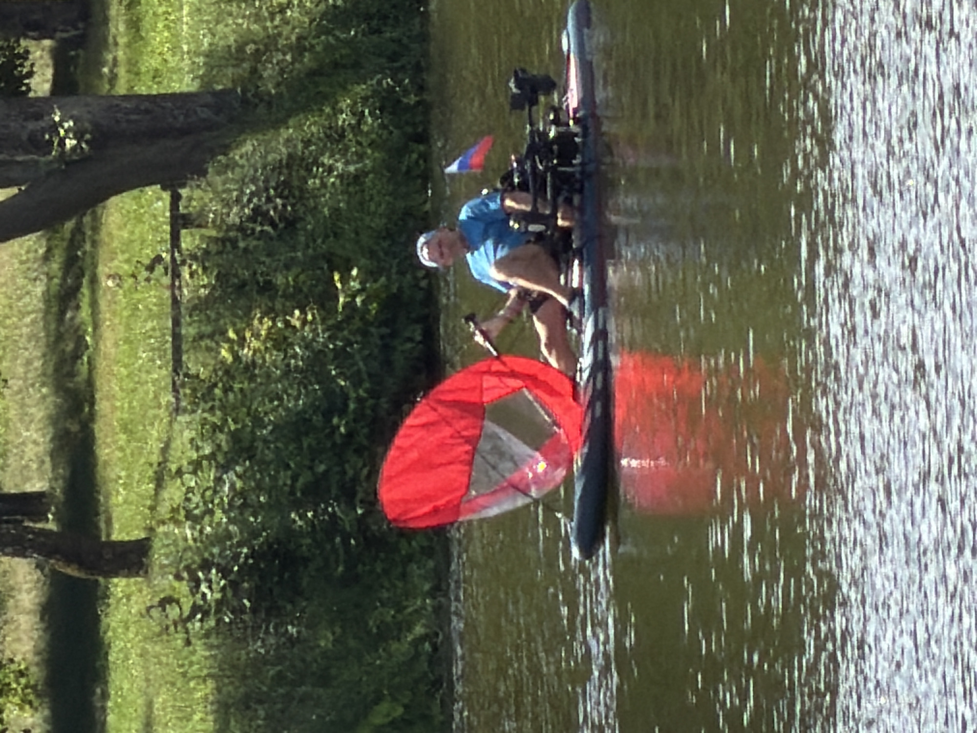 A man is seated on a paddleboard on a body of water. He is wearing a blue t-shirt and a white cap. The paddleboard has a large, red canopy attached to it, and there is some electronic equipment behind the man. A small Russian flag is also mounted on the back of the paddleboard. The water is rippled, and the background consists of green trees and foliage. The scene appears to be a calm, outdoor activity.