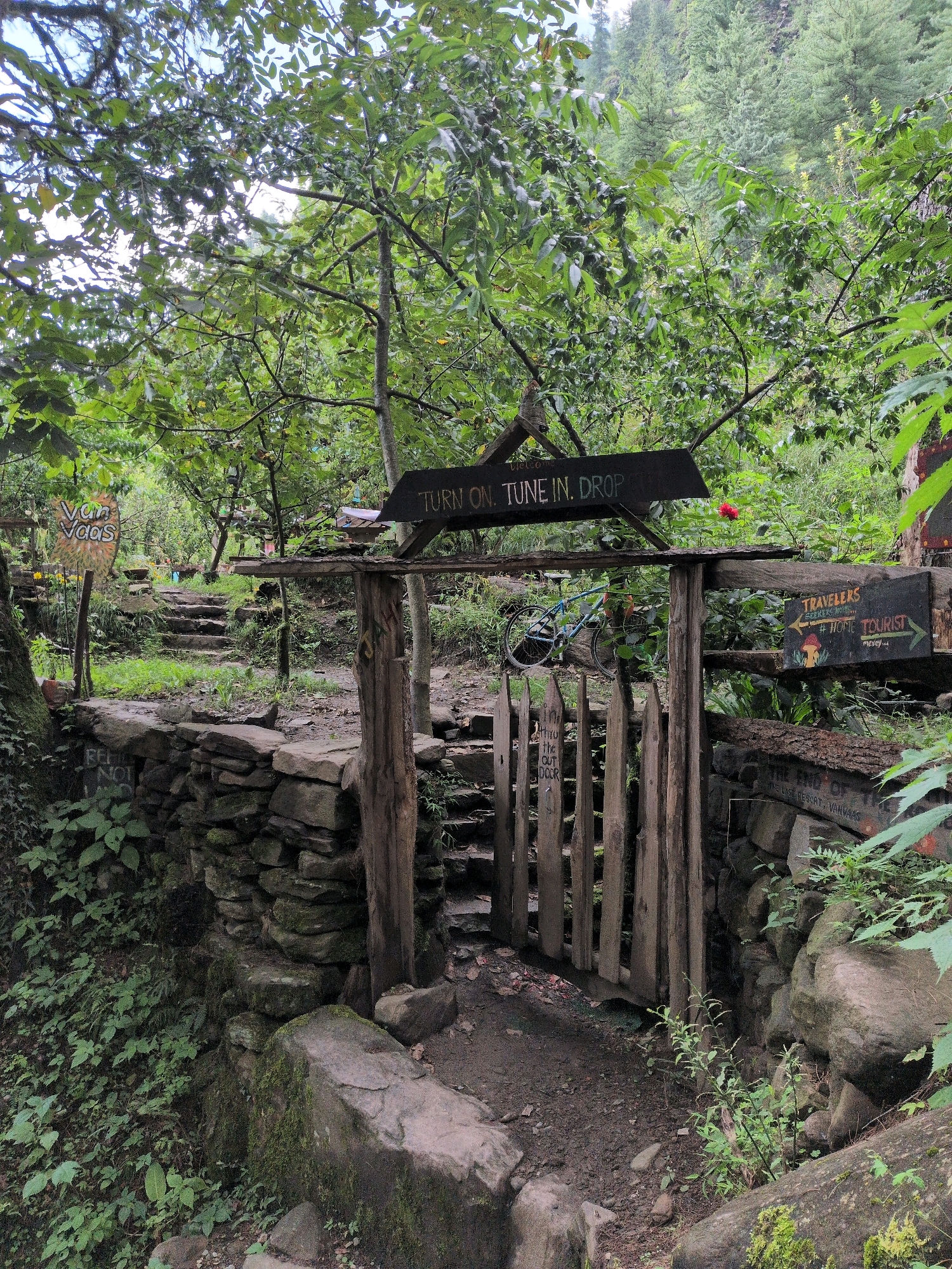 The image depicts an outdoor scene in Dashal, India, featuring a rustic wooden gate and surrounding natural elements. A stone wall, partially covered in moss, runs along the left side of the frame, with lush green foliage growing abundantly. Beyond the wall, a stone pathway ascends into a wooded area.
The gate itself is constructed from rough-hewn wooden planks and stands open, revealing a path leading further into the greenery. Above the gate, a dark wooden sign with white lettering reads "TURN ON TUNE IN. DROP." To the right of the gate, another sign with the word "TRAVELERS" is visible, along with directional arrows and what appears to be a mushroom illustration. A bicycle with a light blue frame is leaning against a log within the property.
In the background, the dense forest canopy dominates the scene, with tall trees and green foliage creating a natural, somewhat overgrown environment. The overall impression is one of a secluded, natural retreat, possibly a campsite or a rural guest house, given the signage. The lighting suggests an overcast or shaded day.