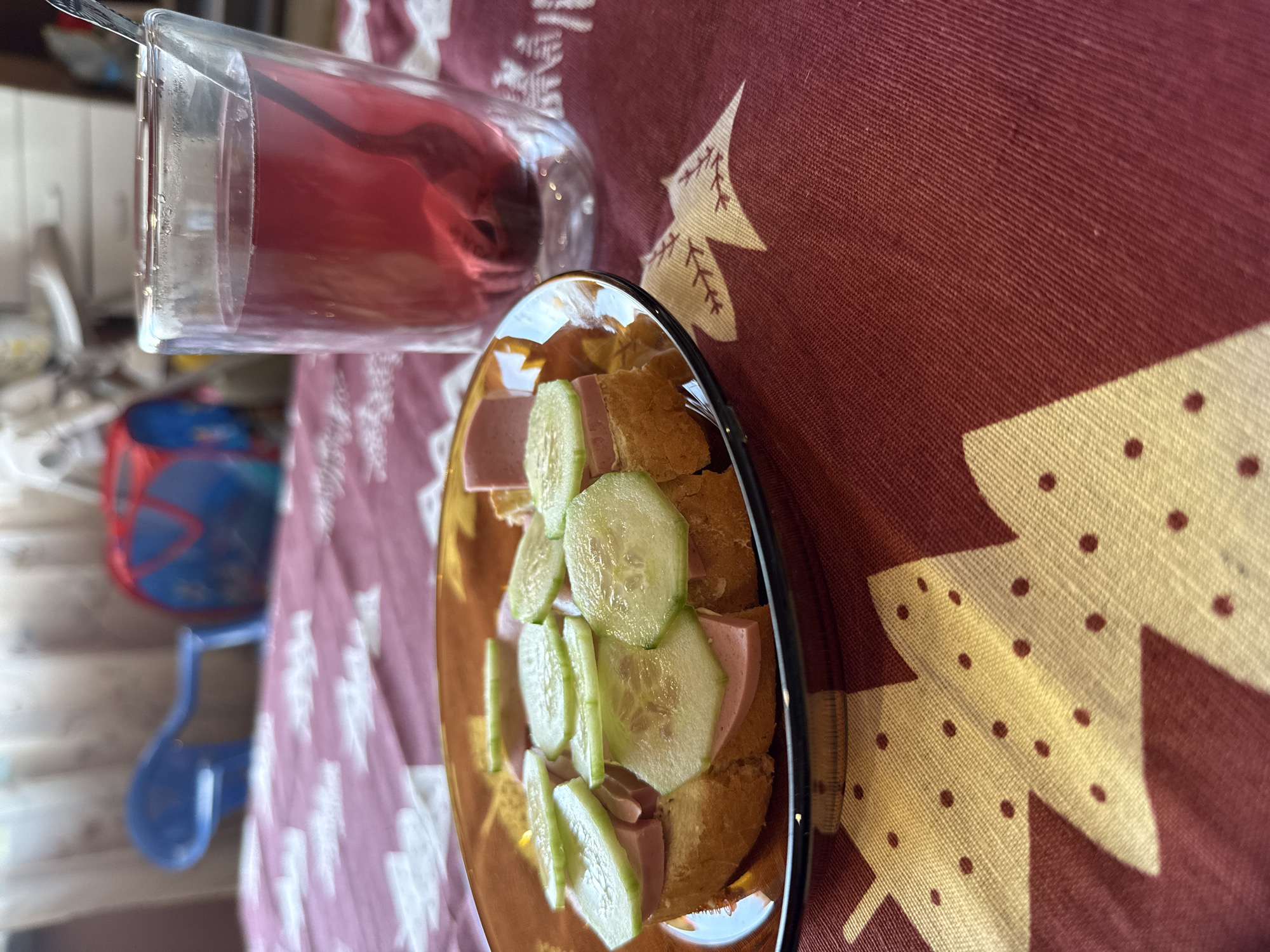 A plate with a sandwich of bread, sausage and cucumber slices rests on a table with a maroon tablecloth decorated with gold Christmas trees. A glass of red liquid is next to the plate. The scene appears to be indoors, and based on the general context and other objects visible in the background, is likely a residential setting. The location is Sofia, Bulgaria.