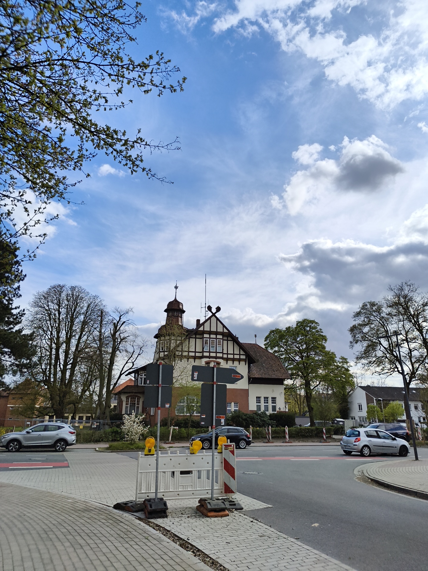 The image depicts a street scene in Diepholz, Germany, featuring a distinctive half-timbered building on the left side of the frame. The building has a tower and appears to be a historical landmark or a building of some significance. The sky is partly cloudy. A street runs in front of the building with several cars visible. There is construction underway as evidenced by the white and red barrier in the foreground. The environment suggests a residential or suburban area within the city.