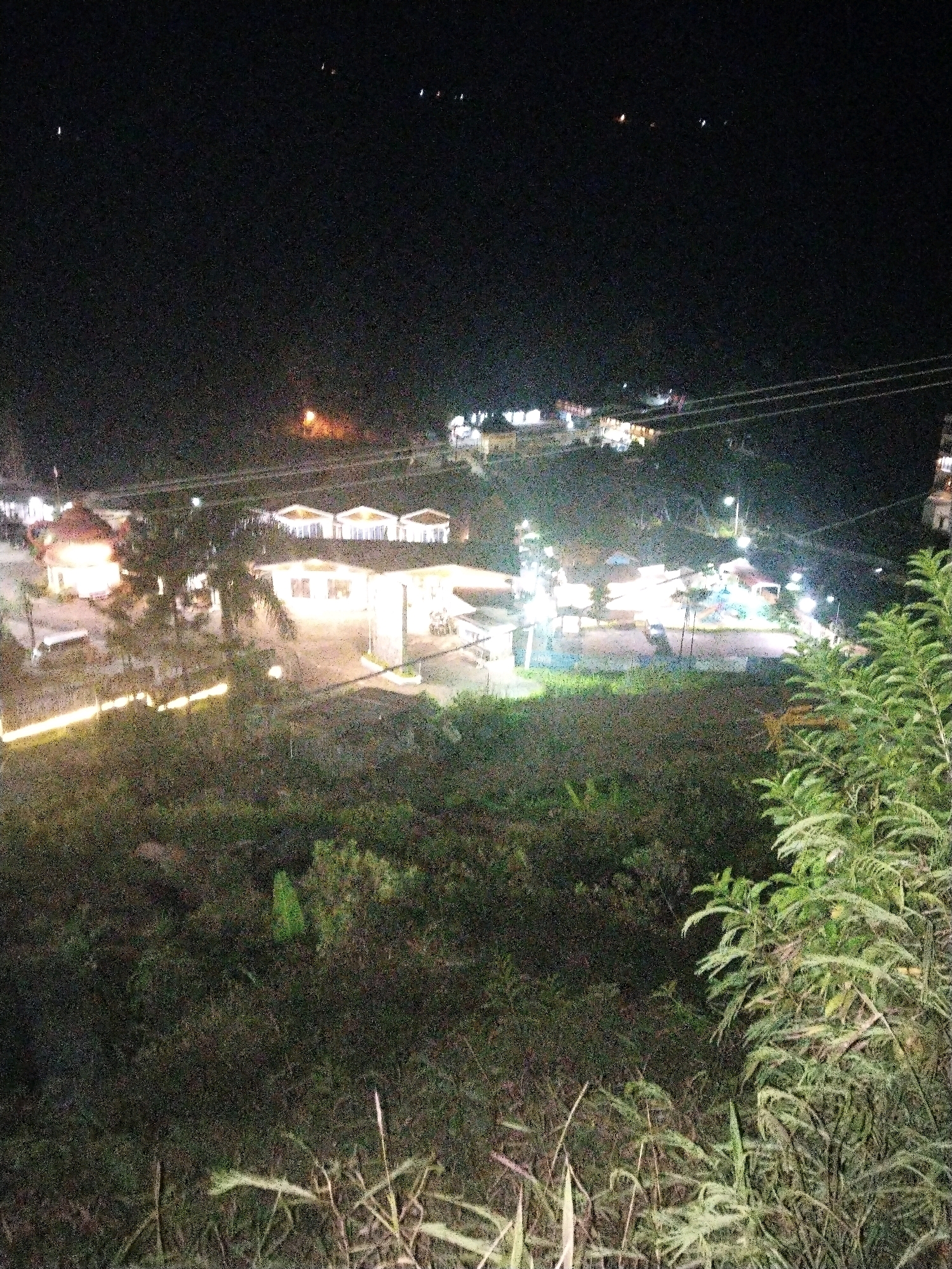 This image is a nighttime aerial view of a well-lit resort or housing complex in the city of Rembul, Indonesia. The resort features several distinct buildings, possibly bungalows or villas, arranged in a row and illuminated with warm, artificial lighting. A central area appears to be a communal space, possibly a restaurant or gathering area, also brightly lit. In the foreground, dense vegetation, including trees and bushes, covers a hillside, obscuring the lower portion of the scene. The sky is dark, with a few distant lights visible, suggesting a clear night. Power lines are also faintly visible spanning across the scene. There are no people visible in the image, and the overall impression is one of a peaceful, illuminated settlement against a dark, natural backdrop.