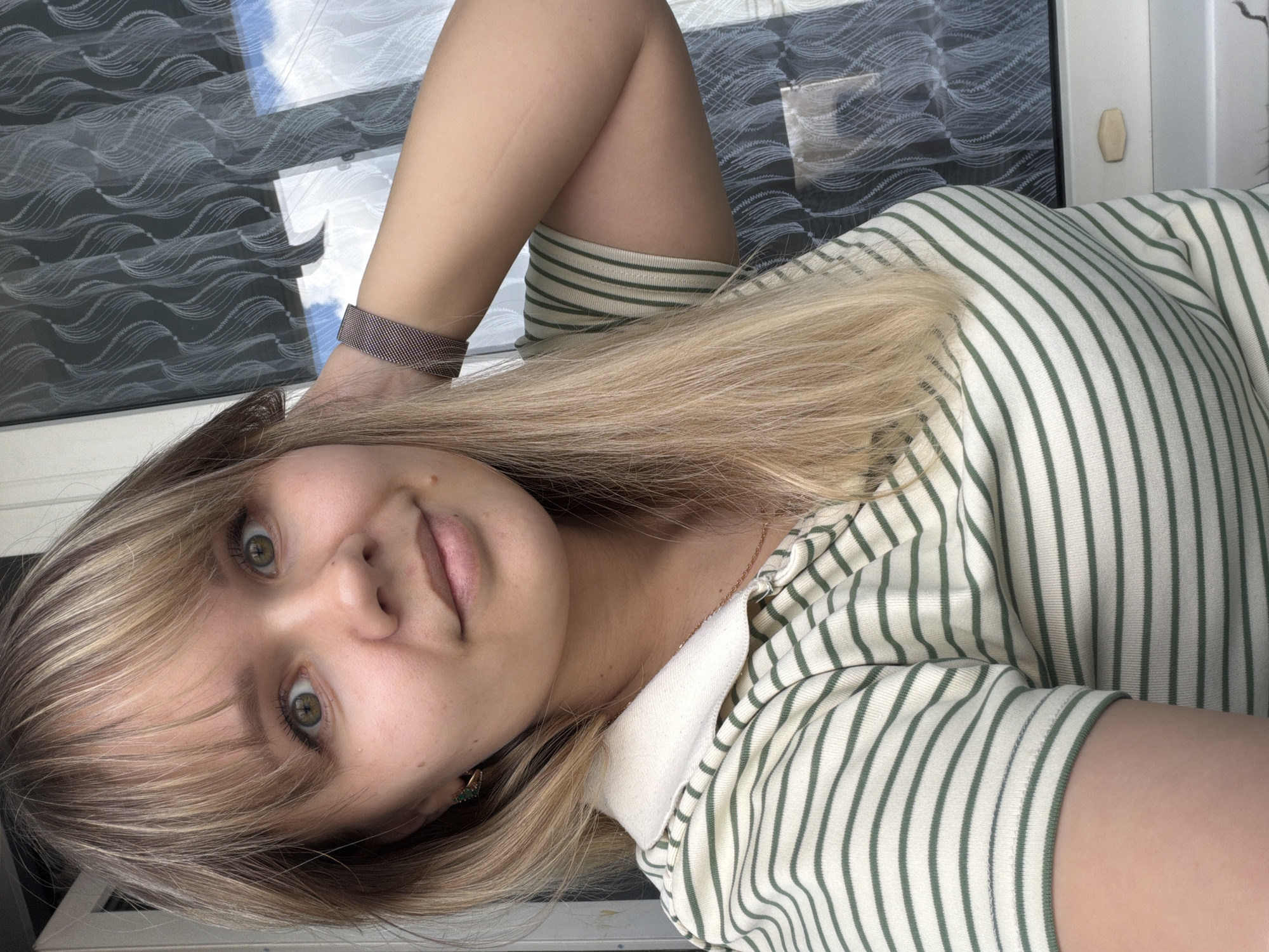 A young woman with light blonde hair and bangs is the central subject of this selfie, which is rotated 90 degrees counter-clockwise. She is looking directly at the camera with wide, greenish-brown eyes and a subtle, closed-mouth smile. Her long, straight hair falls around her face and across her left shoulder and arm.
She is wearing a cream-colored top with thin, olive green horizontal stripes, which appears to have a collar, possibly indicating a polo style. On her left wrist, she wears a dark, possibly rose gold or bronze-toned, mesh-link watch band. A small, green earring is visible in her right ear, and a thin, light-colored (possibly rose gold) necklace chain can be seen against her skin.
The background features a window with a patterned blind or curtain displaying a wavy, almost abstract design in dark grey or black on a lighter background. Through the window, blue sky with white clouds is visible, and the window reflects some light, suggesting bright daytime. A part of a white wall or door frame is also visible, with a small, light-colored, oval-shaped object attached to it. The lighting is natural and bright. No visible text is present in the image.