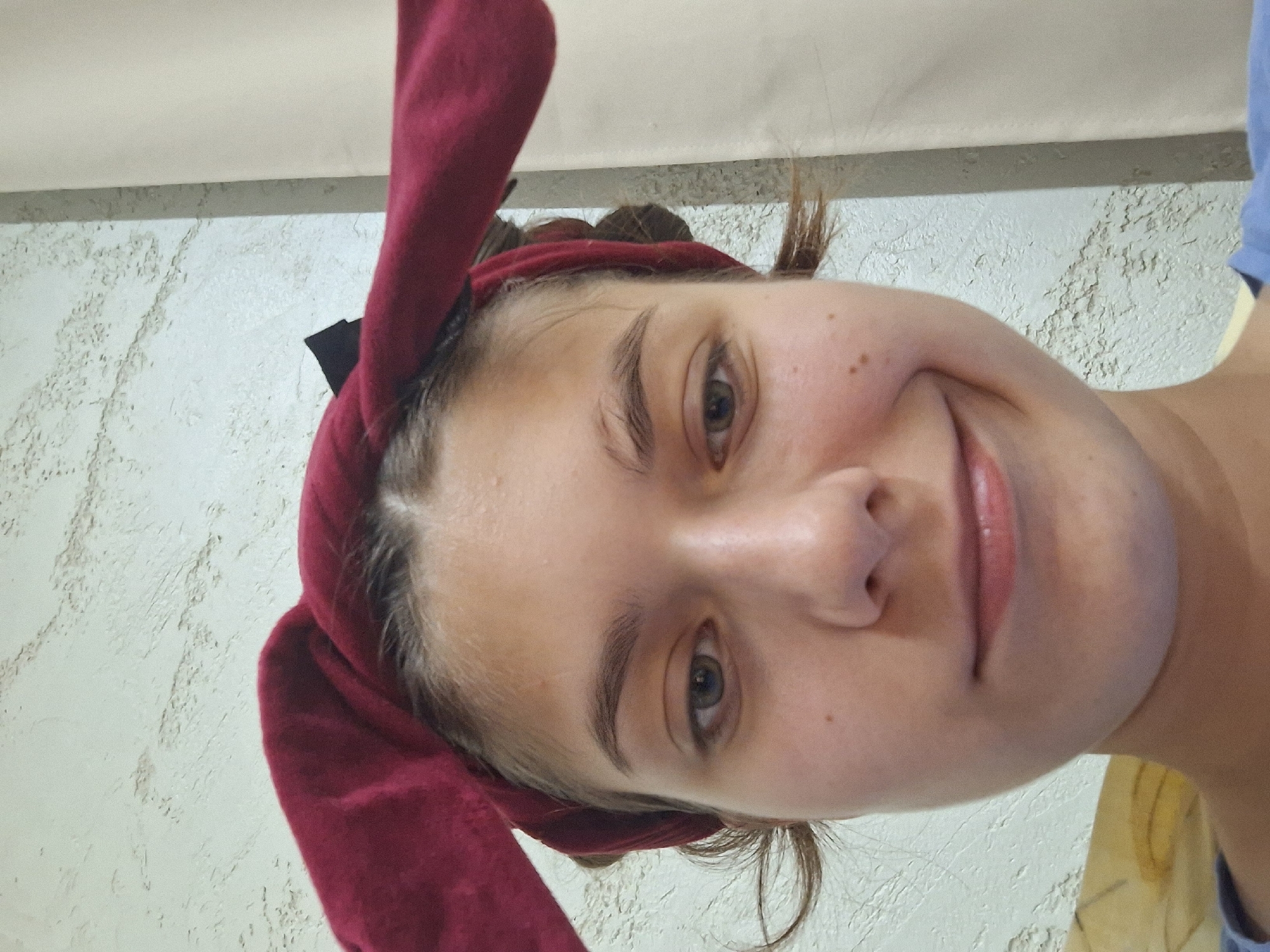 A young person is pictured in a close-up selfie, wearing a burgundy headband with ear-like protrusions. The individual has light skin, brown eyebrows, and green eyes. Their mouth is turned up in a slight smile. The background appears to be a textured wall. The image was taken in Panagyurishte, Bulgaria.