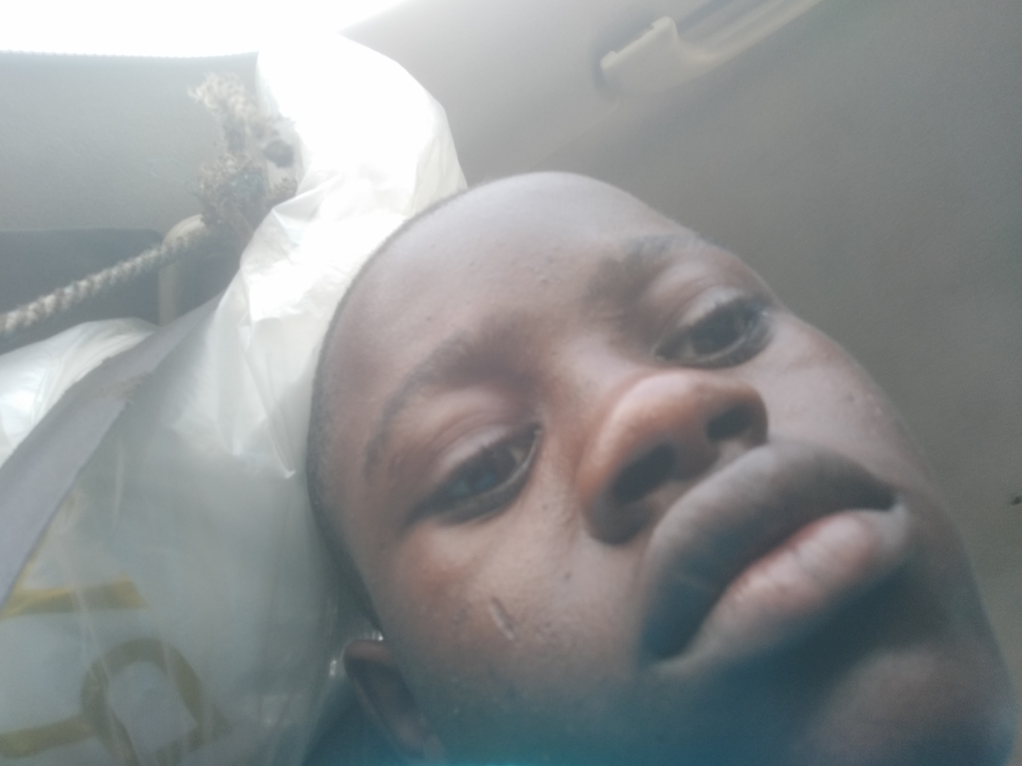 A close-up, low-angle photograph shows a dark-skinned individual's face, potentially male, with their head resting on a white plastic bag inside what appears to be a vehicle. The individual's features are prominent, including their eyes, nose, and lips. A scar is visible on the cheek. A rope-like object is visible near the plastic bag. The interior of the vehicle is partially visible, including a portion of the ceiling. The location in the city of Ilishan, Nigeria is not verifiable.