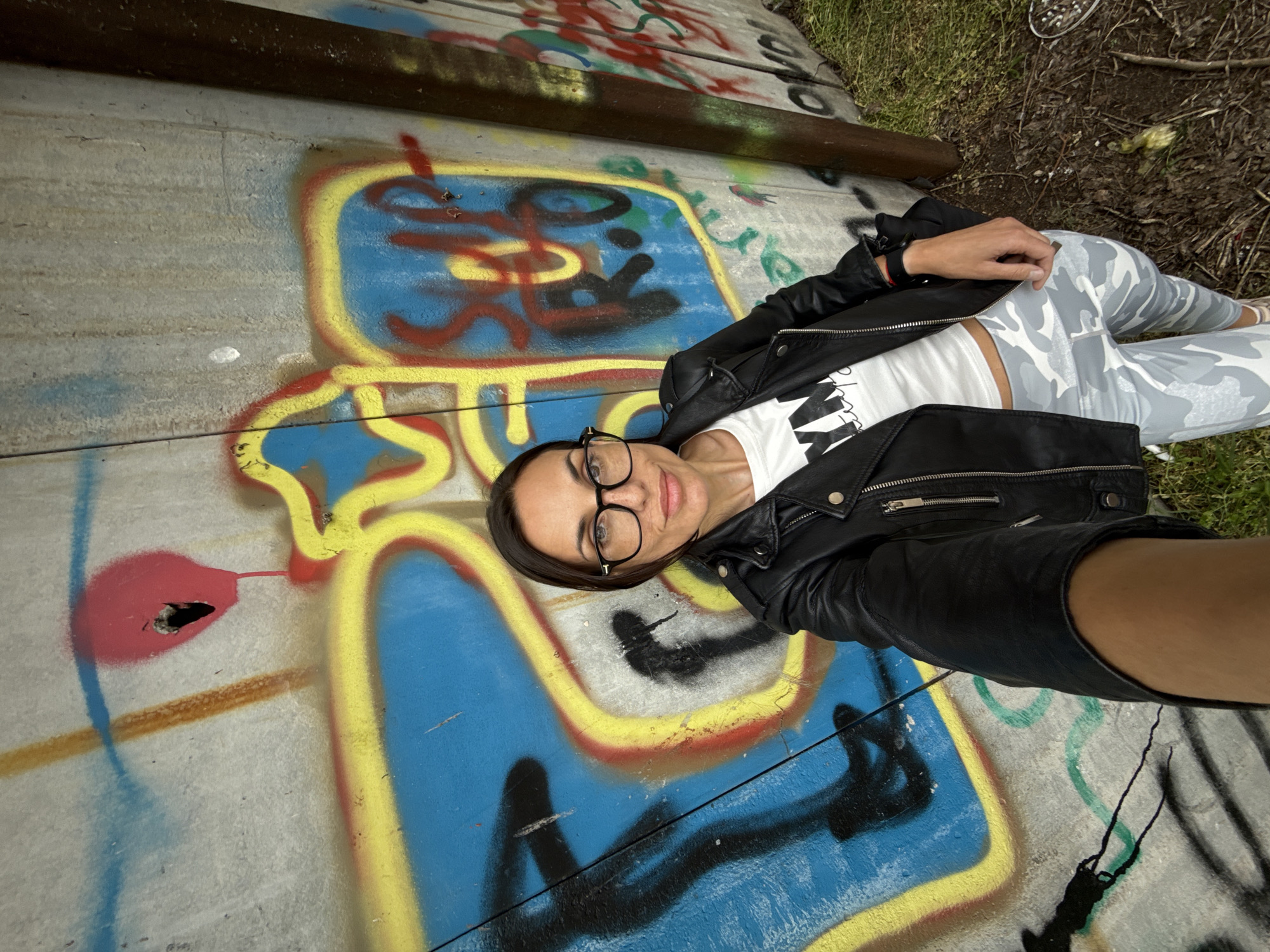 A woman is depicted in a full-frame shot, lying or leaning on a graffiti-covered concrete surface, looking directly at the camera with a relaxed expression and a slight smile. She wears black-rimmed glasses, a black leather jacket, a white t-shirt with the stylized black letters "AW" visible (likely part of a larger word), and light-colored camouflage-patterned leggings. Her dark hair is parted to the side.
The concrete surface beneath her is extensively covered in vibrant, multi-colored graffiti, featuring large blue and yellow outlines, red splashes, and black and green tags forming abstract shapes and illegible letters. A dark, rusty metal rail or pipe runs across the upper part of the concrete. Beyond the concrete, patches of natural ground with dirt and sparse vegetation are visible in the top right and bottom right corners. The lighting appears soft and even, suggesting daytime, possibly under an overcast sky. The setting is an outdoor urban or industrial environment, such as a wall or underpass, located in Pleven, Bulgaria.