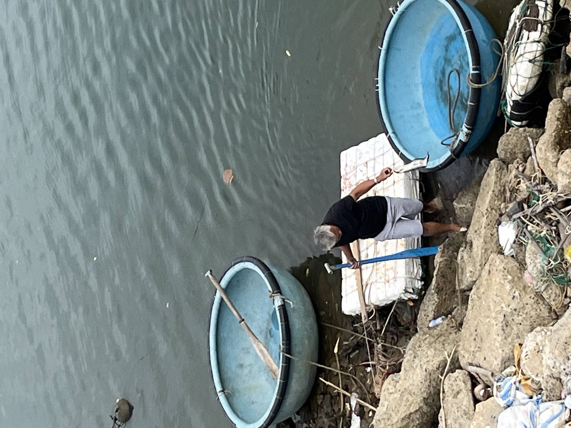 The image depicts a man in a black t-shirt and gray shorts standing on the rocky shore of a body of water, likely in Nha Trang, Vietnam. He is holding a small fish in his left hand, and in his right hand, he is holding a blue paddle. In front of him is a circular blue boat, possibly a coracle, with a rope coiled inside. To his left, further from the shore, is another similar blue boat with a wooden paddle inside. The water is calm and appears to be a dark green color. The shore is littered with rocks, debris, and fishing nets. A white raft-like object is partially submerged in the water near the man. The scene suggests a fishing activity or preparation for it.
