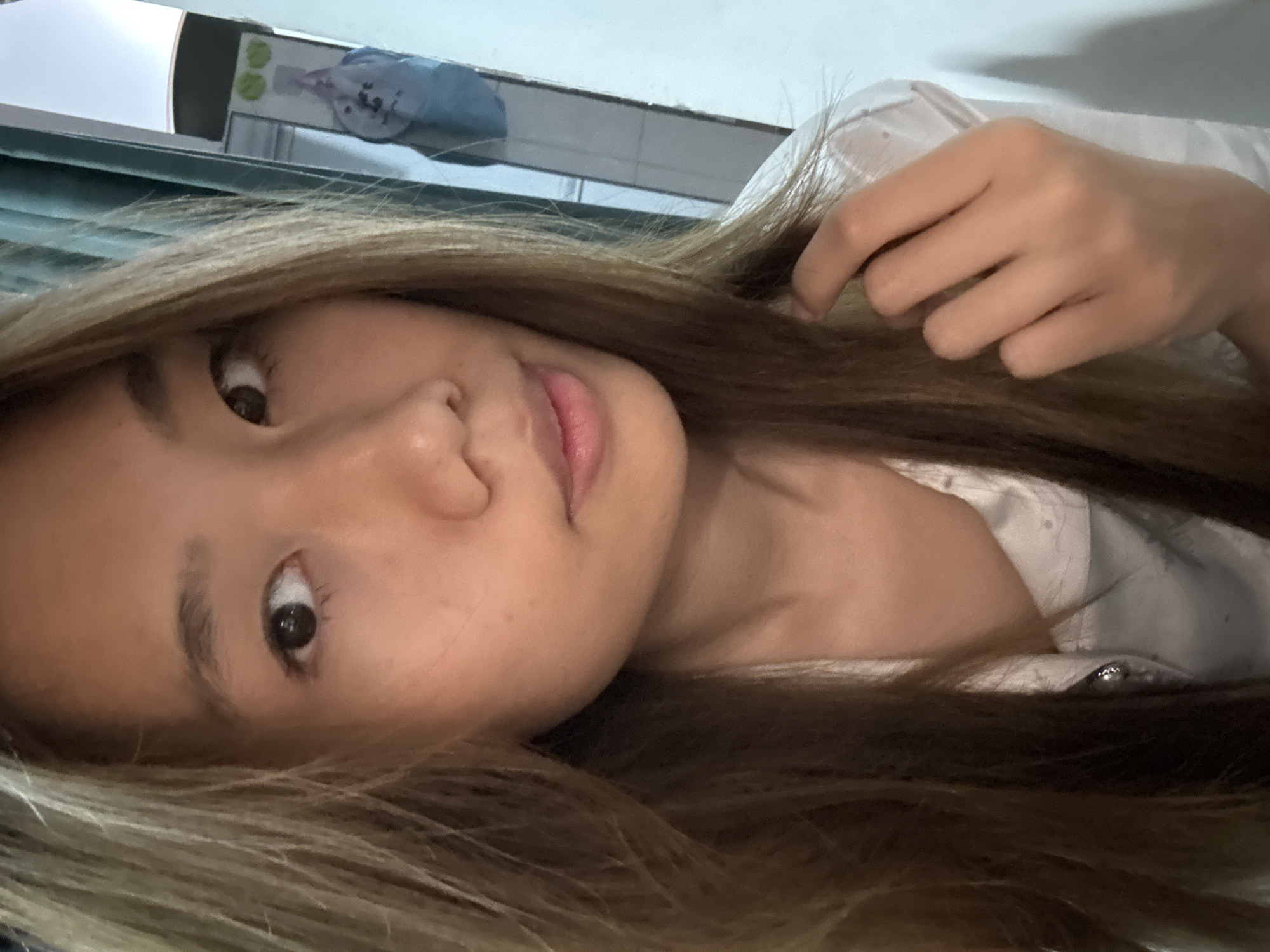 A close-up, rotated selfie shows a person with light brown and blonde-highlighted hair, light skin, and brown eyes. The person is wearing a white button-down shirt. A hand is visible near the person's hair. In the background, a white surface with a television screen is partially visible. There is no indication of the location being Nan'an District, China, or any specific city.