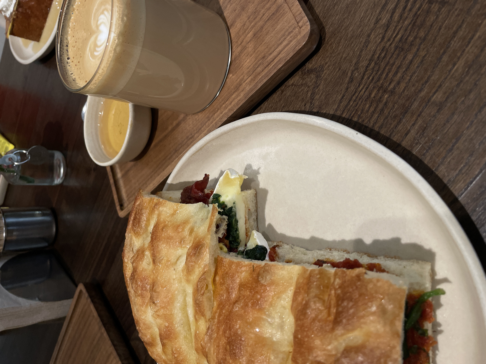 The image captures a meal scene likely in Milan, Italy, given the style and presentation of the food and drink. On a wooden table, a plate features a sandwich made with focaccia bread, revealing fillings such as tomatoes, spinach, and soft white cheese. Adjacent to the plate is a glass of latte and a small white bowl with honey. In the periphery, a piece of cake on a plate is visible. The overall setting indicates a café or restaurant environment.