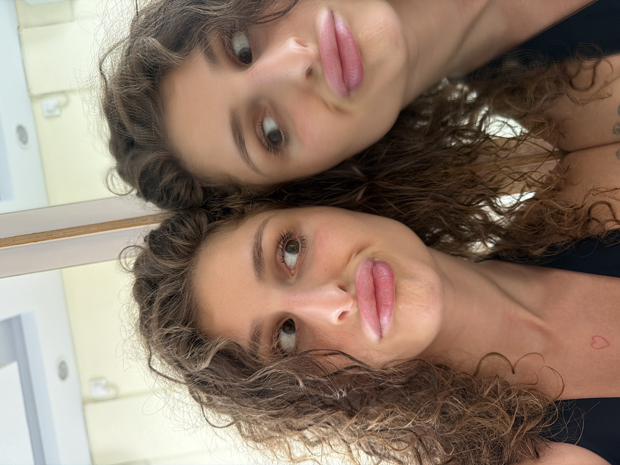 A young woman with curly brown hair is taking a mirror selfie. Her face is partially obscured by her hair, and she is making a duck face. The background is a brightly lit room with a white ceiling and cream-colored walls. The woman is wearing a black top. There is a small red heart tattoo visible on her chest. The reflection in the mirror shows another part of the woman's face and hair, as well as the surrounding room. The image appears to have been taken in a bathroom or bedroom.