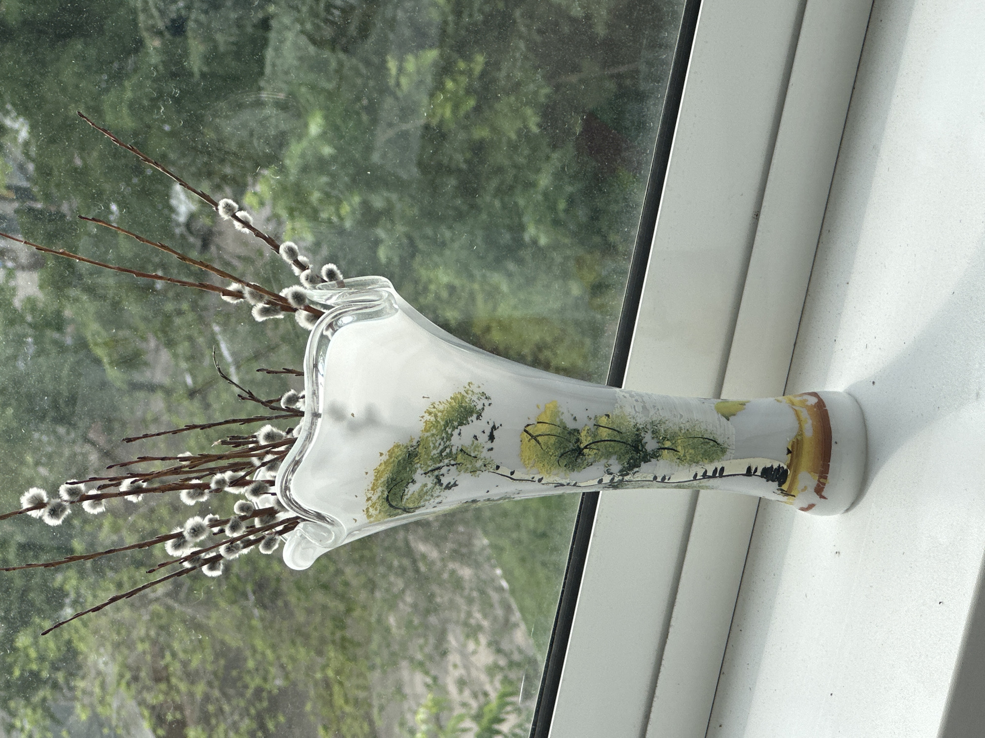 A tall, slender glass vase with a flared opening sits on a white windowsill. The vase is predominantly white, adorned with a painted scene of stylized trees with yellow and green foliage and black branches. A band of brown and yellow paint encircles the base of the vase. Inside the vase are several branches of pussy willows, their fuzzy catkins catching the light.
The vase is positioned in front of a window that looks out onto a blurred backdrop of green foliage, suggesting a natural setting. The windowpane is slightly dusty. The lighting indicates it could be daytime, with natural light filtering through the window. The overall scene has a calm and natural feel, possibly evoking a sense of spring due to the pussy willows. The windowsill and surrounding window frame are white, with subtle texture suggesting they are painted or made of a similar material. No people are visible in the image, and no text is present. The context of Birobidzhan, Russia, places this scene in a specific geographic location.