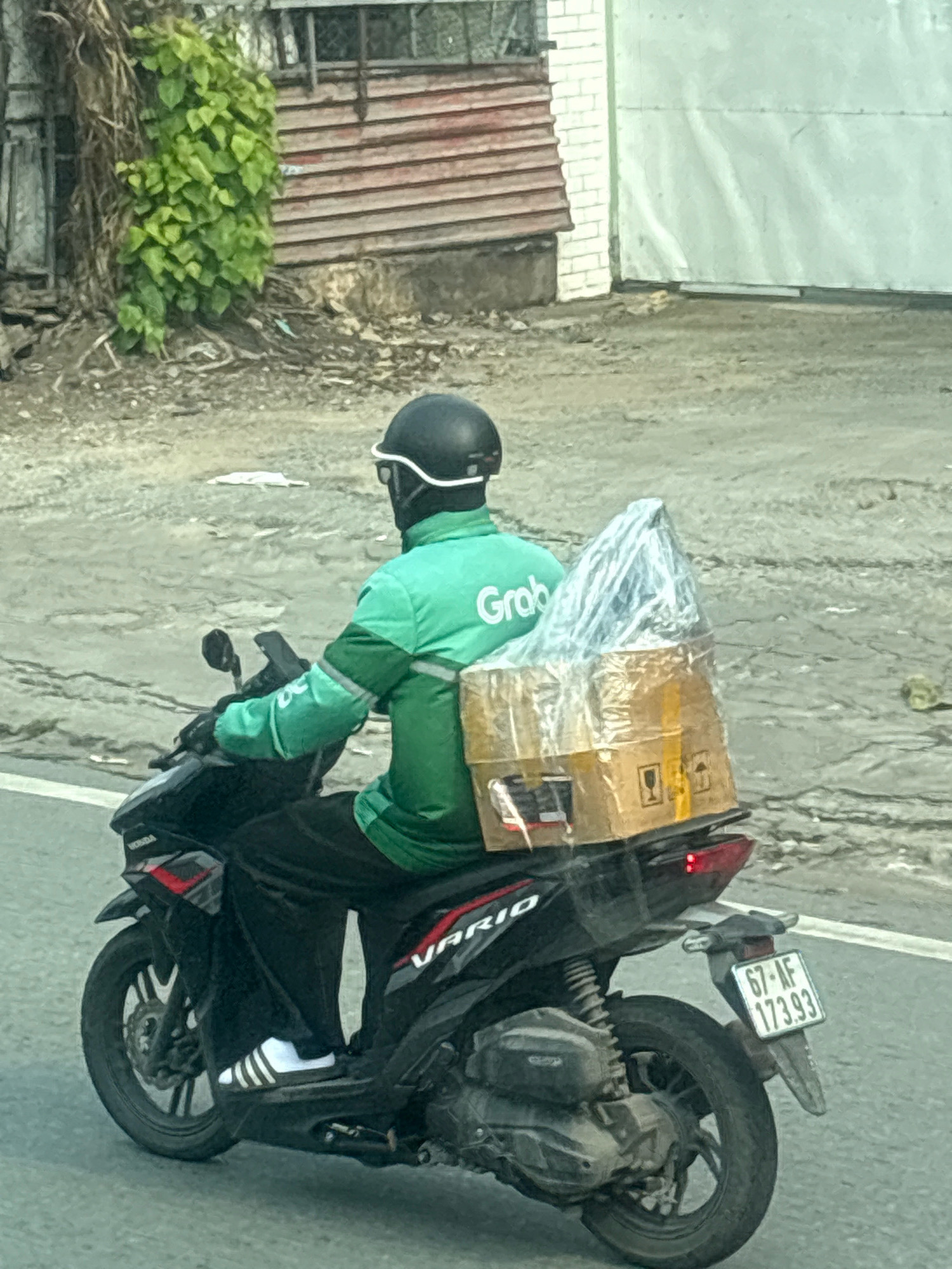 A person wearing a green Grab delivery jacket and a black helmet is riding a black motorcycle with a license plate reading "67-AF 173.93" on a paved road in Ho Chi Minh City, Vietnam. The motorcycle has a large cardboard box and a plastic-wrapped item strapped to the back. The background shows a building with red wooden siding and green foliage growing on a vine. The road appears to be somewhat rough and cracked. The weather is overcast.