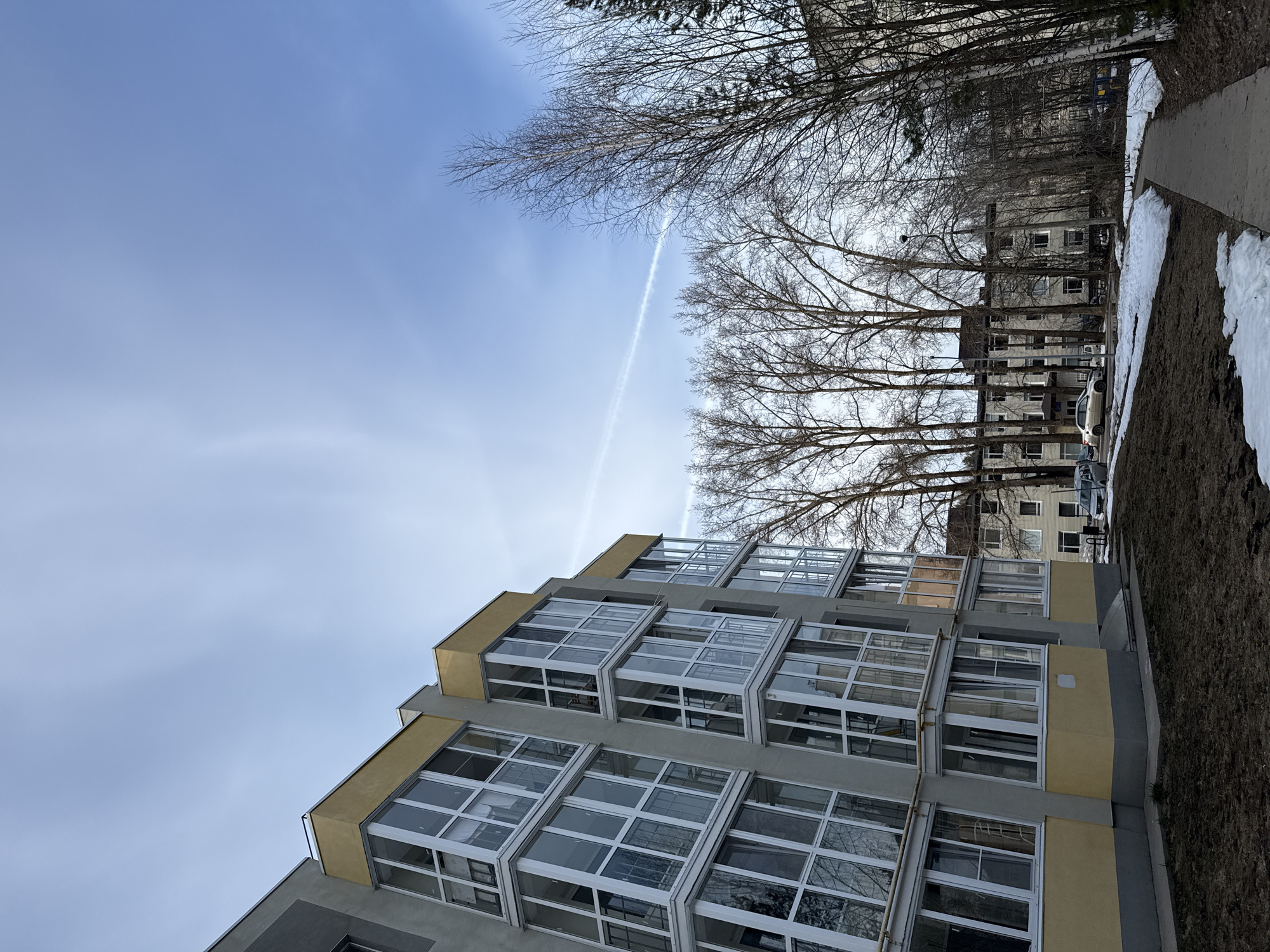 The image shows an outdoor scene in an urban setting, with a modern building featuring many windows taking up a significant portion of the frame. The building is predominantly gray and yellow. Above the building, the sky is visible, with a contrail cutting across it. Bare trees are situated along the upper edge of the scene and in the background, partially obscuring a row of buildings. Patches of snow are visible along the right edge, next to an area of dry grass and a sidewalk. A car is parked next to a building in the background. The scene conveys a sense of early spring or late winter.