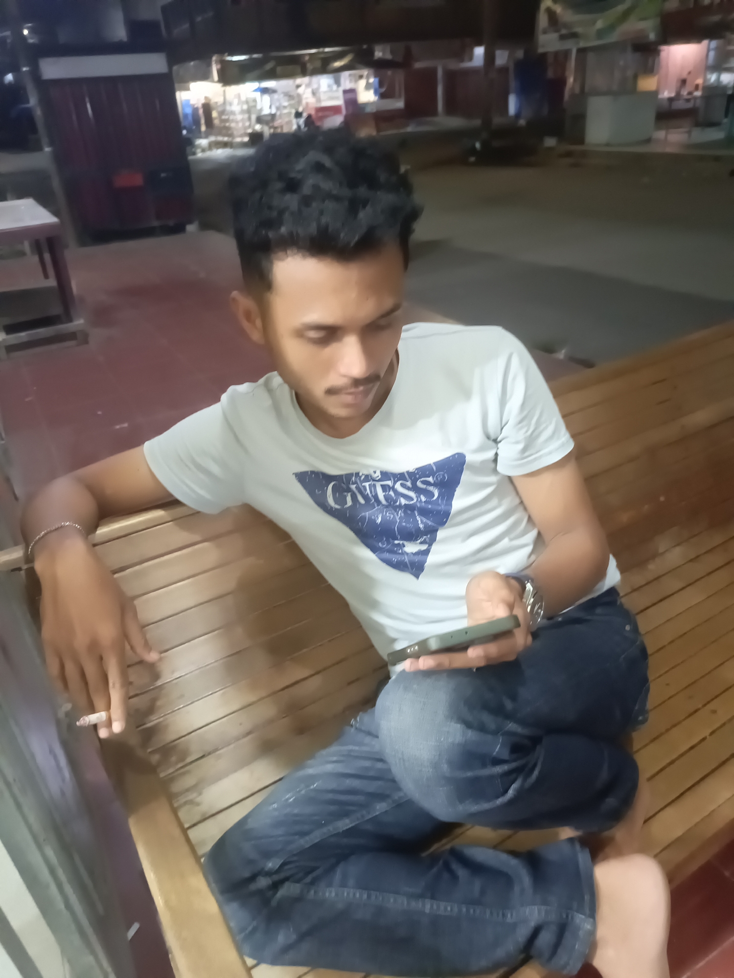 A young man with dark, curly hair and a mustache sits on a wooden bench outdoors at night. He wears a light blue t-shirt with a graphic design and blue jeans. He holds a smartphone in his right hand and a lit cigarette in his left hand, which is resting on the bench. The bench is situated in an area with a red floor, suggesting it might be a public space such as a park or a rest area. In the background, there are buildings with illuminated signs, indicative of a commercial or street scene in the city of Lhokseumawe, Indonesia.