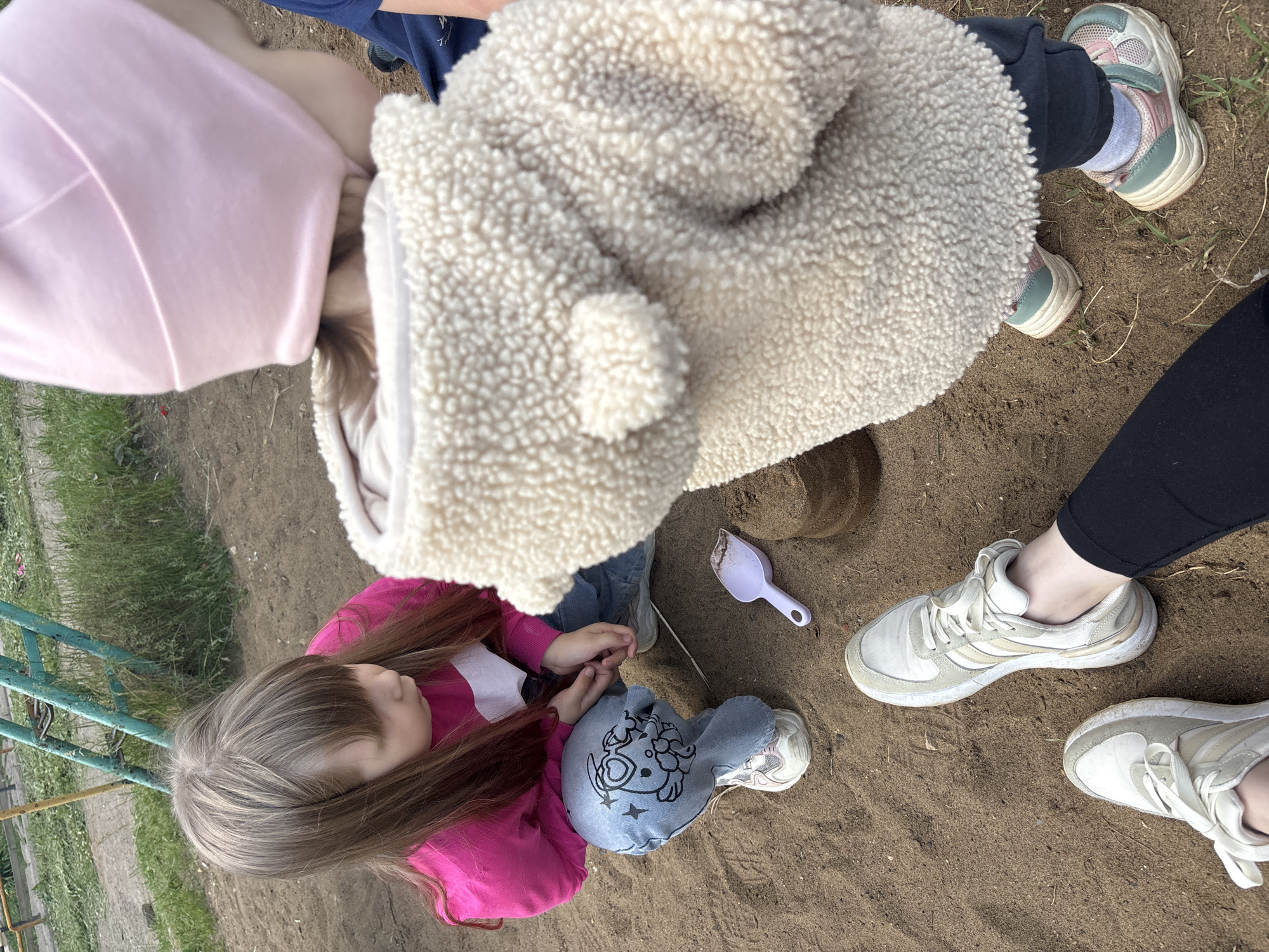 An overhead photograph captures a group of people, primarily children, playing in a sandy area, likely an outdoor playground in Chaikovsky, Russia. The perspective looks down onto the scene.
In the center-left, a child with long, light brown hair sits on the sand, wearing a bright pink long-sleeved shirt and light blue jeans featuring a black cartoon graphic of a character, possibly a dog or sheep, surrounded by stars. This child's hands are clasped together.
To the right of the sitting child, another child is partially visible, wearing a cream-colored, fluffy, hooded jacket and dark pants, with feet in light-colored sneakers with pink and teal accents. A small, light purple plastic shovel lies in the sand near them, beside a sculpted mound of sand.
In the upper left corner, the back of a head wearing a pink hood or hat is visible. To the far right, the lower legs and feet of an adult or older child are seen, wearing black leggings and white sneakers, standing on the sand.
The ground is predominantly light brown sand. To the left, patches of green grass and a weathered, teal-colored metal structure, characteristic of playground equipment, are visible. The natural lighting suggests it is daytime, and the clothing indicates mild to cool weather. No discernible text is visible on any objects or clothing, apart from the graphic design on the jeans. The overall mood is one of casual outdoor play.