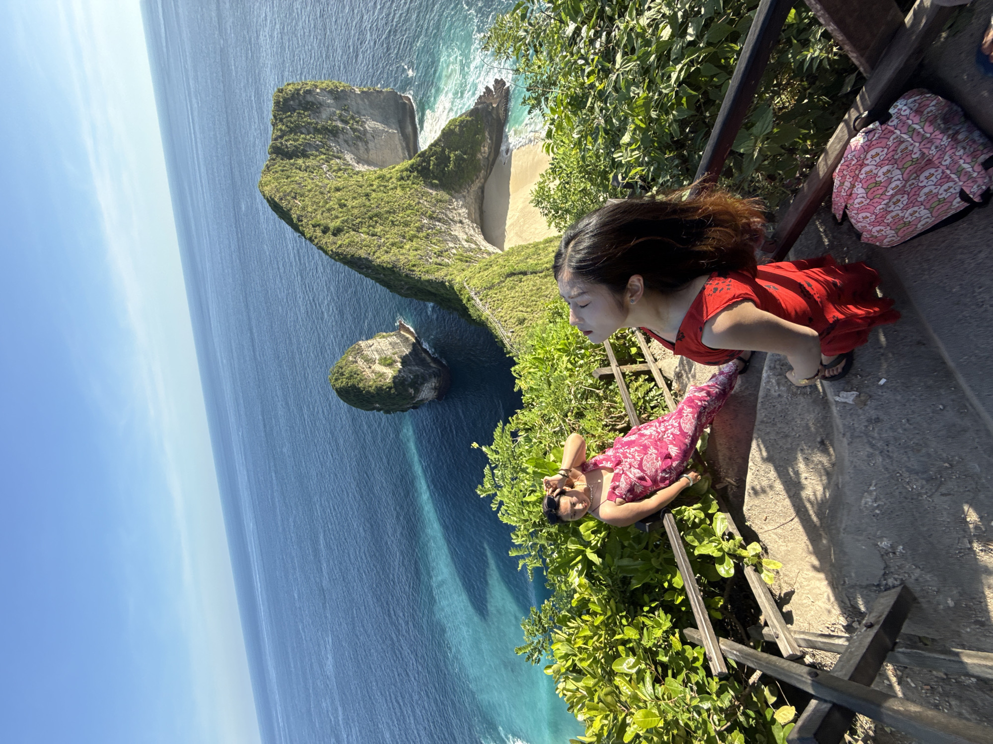 The image captures a scenic view from Nusa Penida, Indonesia. The landscape features a prominent, rocky coastline, with green vegetation covering the cliffs and turquoise blue waters of the ocean stretching to the horizon. Two women are positioned on a set of concrete steps, seemingly part of a pathway or viewpoint. One woman wears a red dress and stands looking towards the cliffside and ocean. The other woman is lying down, wearing a pink patterned dress, with her head resting near the foliage. A pink patterned backpack is visible on the steps. The scene is illuminated by bright daylight.