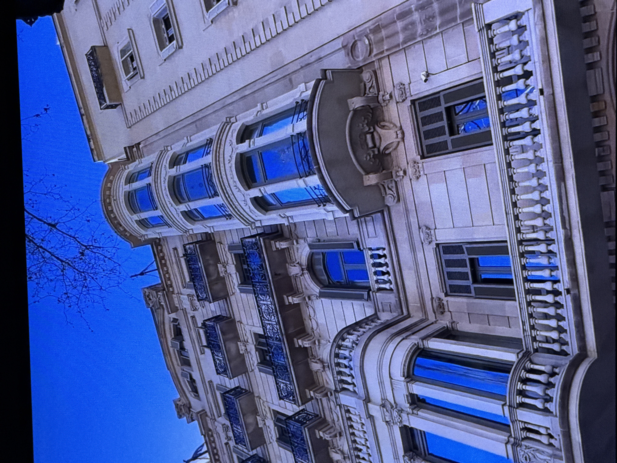 The media file shows an upward-angled shot of a pale stone building with ornate architectural details. The building has multiple stories, featuring numerous windows, some of which have dark shutters and others with bright blue glass. There are balconies with decorative railings and intricate stonework. A bare tree branch is visible on the left against a clear, vibrant blue sky. The overall impression is of a grand, possibly older, building in an urban setting. There is no visible text. The bright blue sky suggests it is daytime, and the clear conditions indicate fair weather. Given the location context of Dubai, United Arab Emirates, the building's architecture might reflect influences from that region, though its style also has a classical European feel. No people or specific activities are depicted; the focus is on the building's facade and its interaction with the sky and natural light. The image is shot from a low angle, emphasizing the height and grandeur of the structure.
