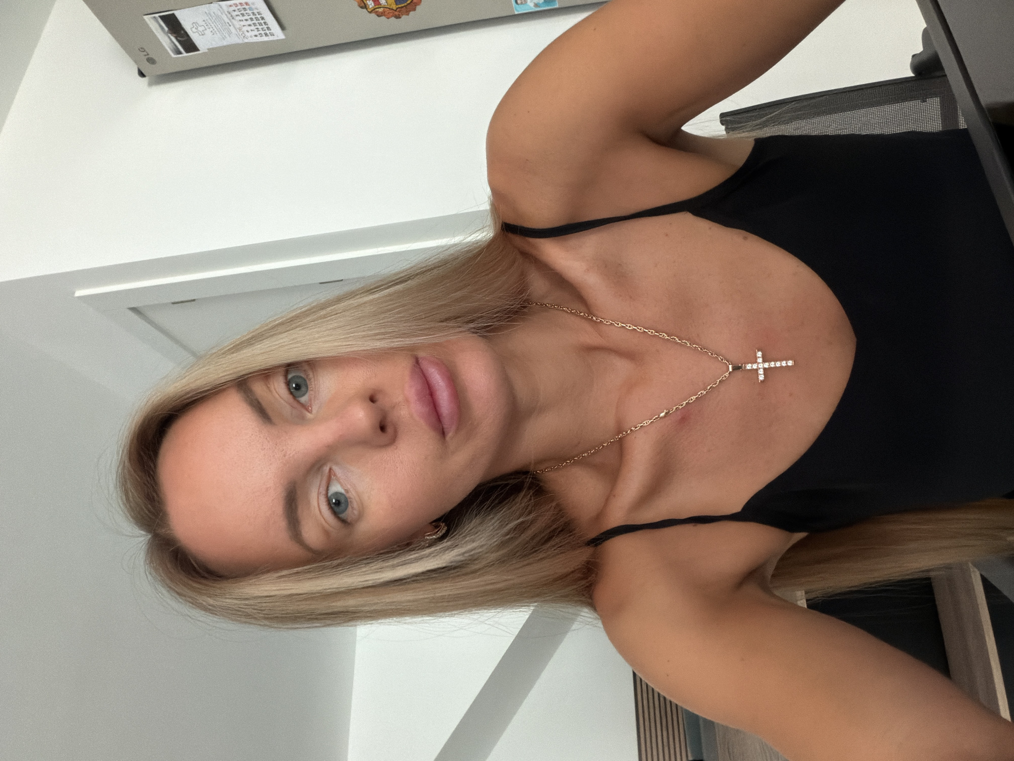 An adult Caucasian female with long blonde hair and blue eyes is taking a selfie indoors. She is wearing a black tank top and a gold chain with a cross pendant. Her lips are slightly parted and she is looking directly at the camera. She is sitting in what appears to be a modern kitchen or dining area, with a white refrigerator in the background. The environment is brightly lit, suggesting daytime. The background also includes a white door and part of a chair. There is no indication of the specific location within the city of Finestrat, Spain, based on the visual information provided.